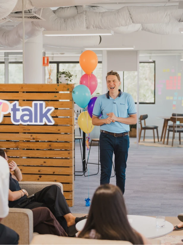 Man giving presentation at office with balloons. Corporate event, "Talk" company logo, smiling. Inside office space, people in attendance.