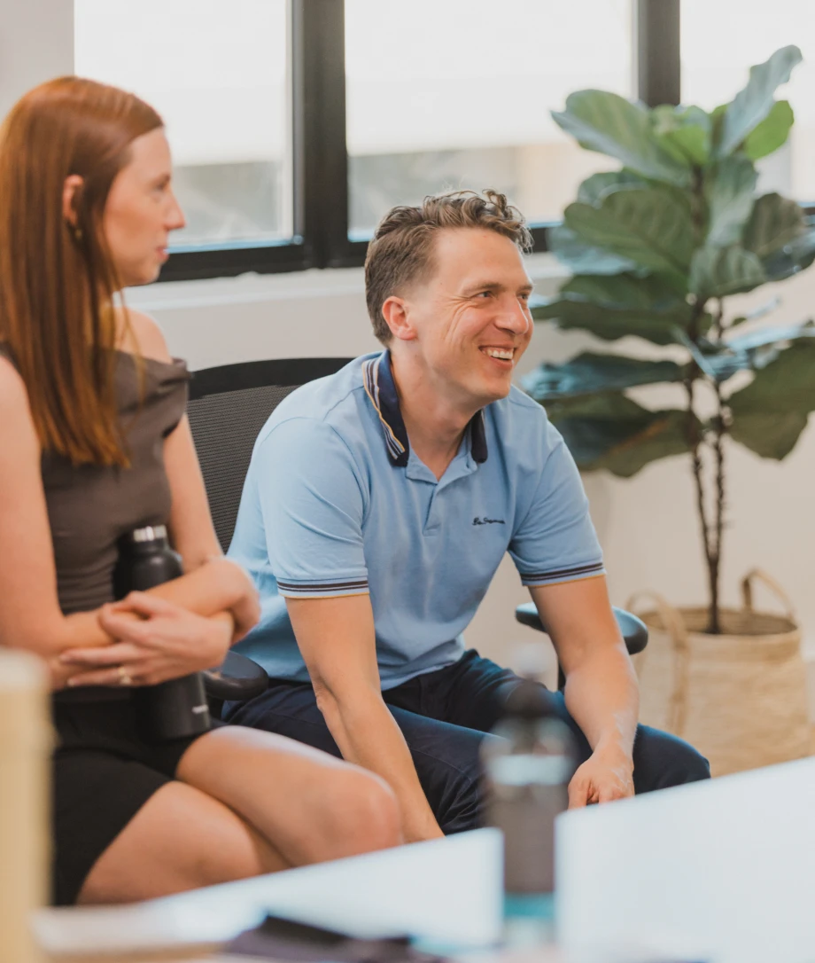 Business meeting in office with two people, man smiling, woman holding water bottle, discussion, collaborative workspace.