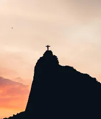 Silhouette of Christ the Redeemer statue atop mountain, silhouetted against sunset sky.
