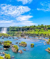 Waterfalls cascading into a river, lush green foliage and a clear blue sky over tropical landscape.