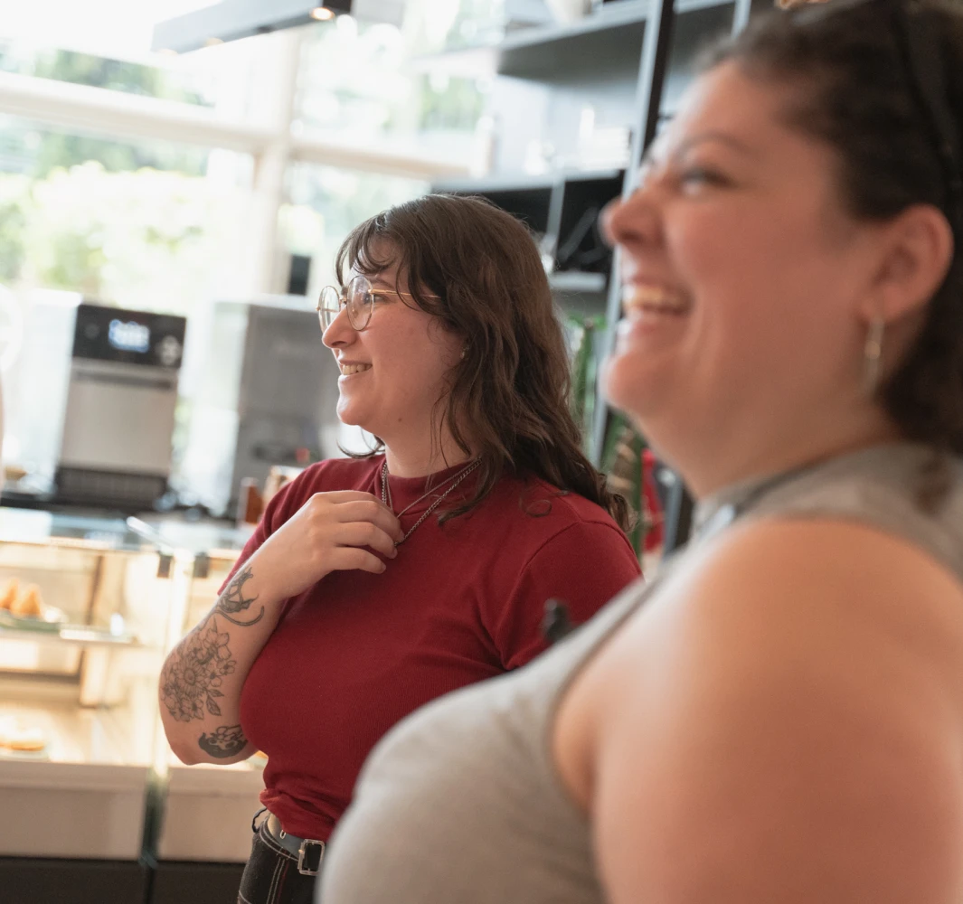 Two women laughing indoors, one with glasses & a tattoo. Cafe or restaurant setting.