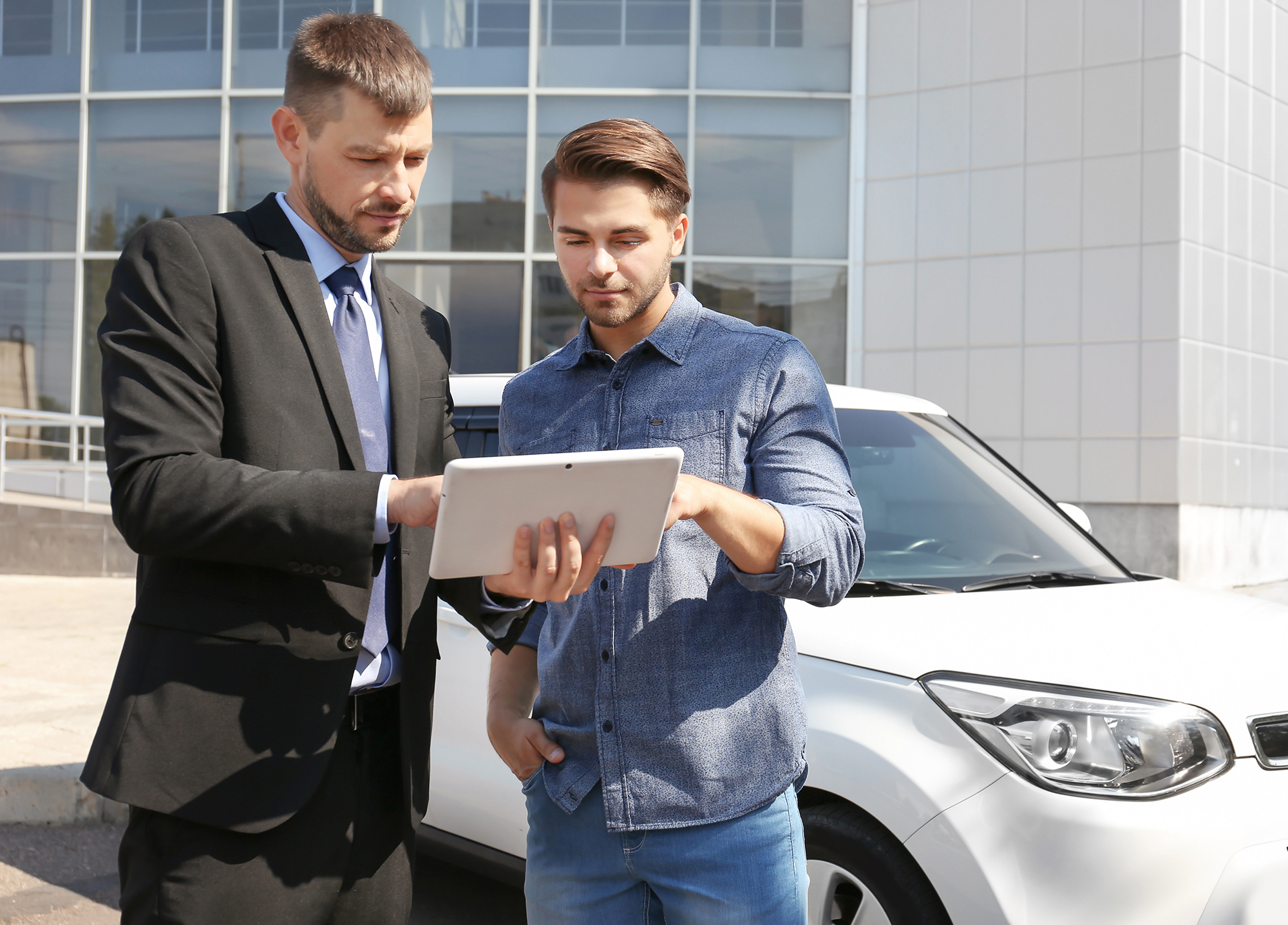 Two men standing by a white car, looking at a tablet, one in a suit and the other in a casual denim shirt.