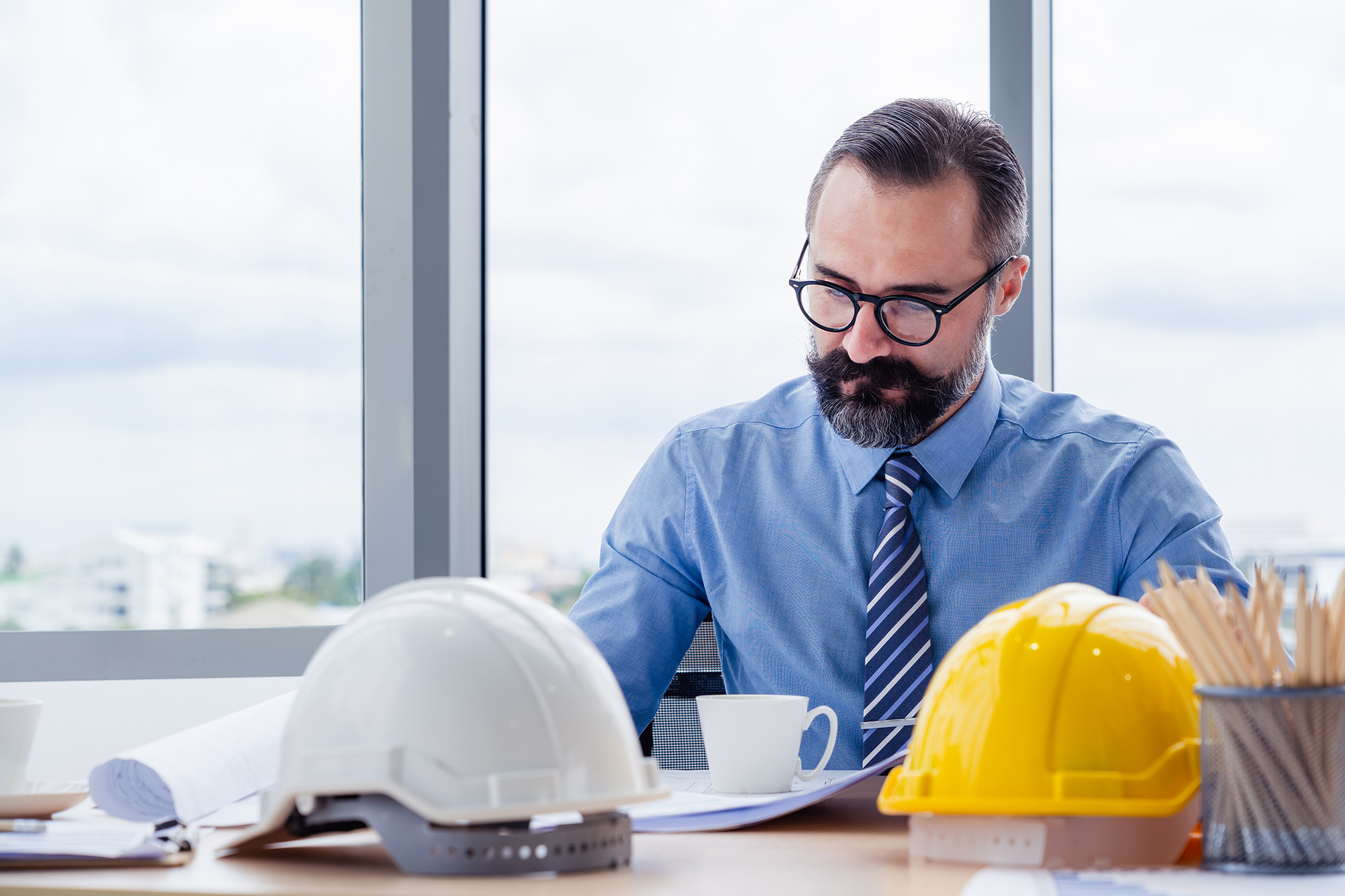Man with glasses and beard in blue shirt and striped tie working at a desk with white and yellow construction helmets and architectural plans.