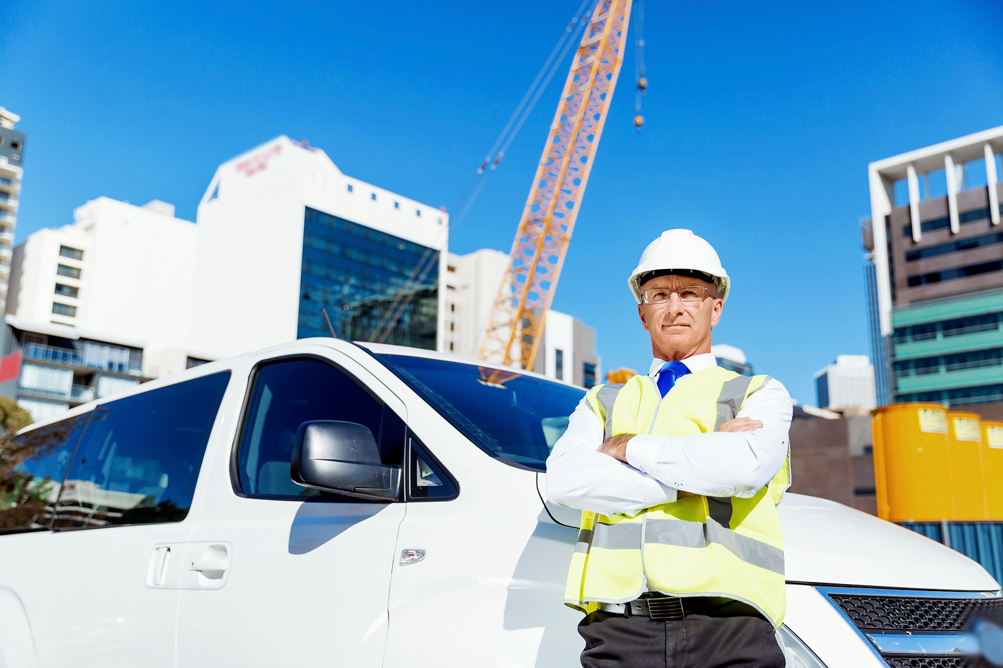 Construction manager wearing a white hard hat and reflective vest standing with arms crossed in front of a white vehicle at a city construction site.
