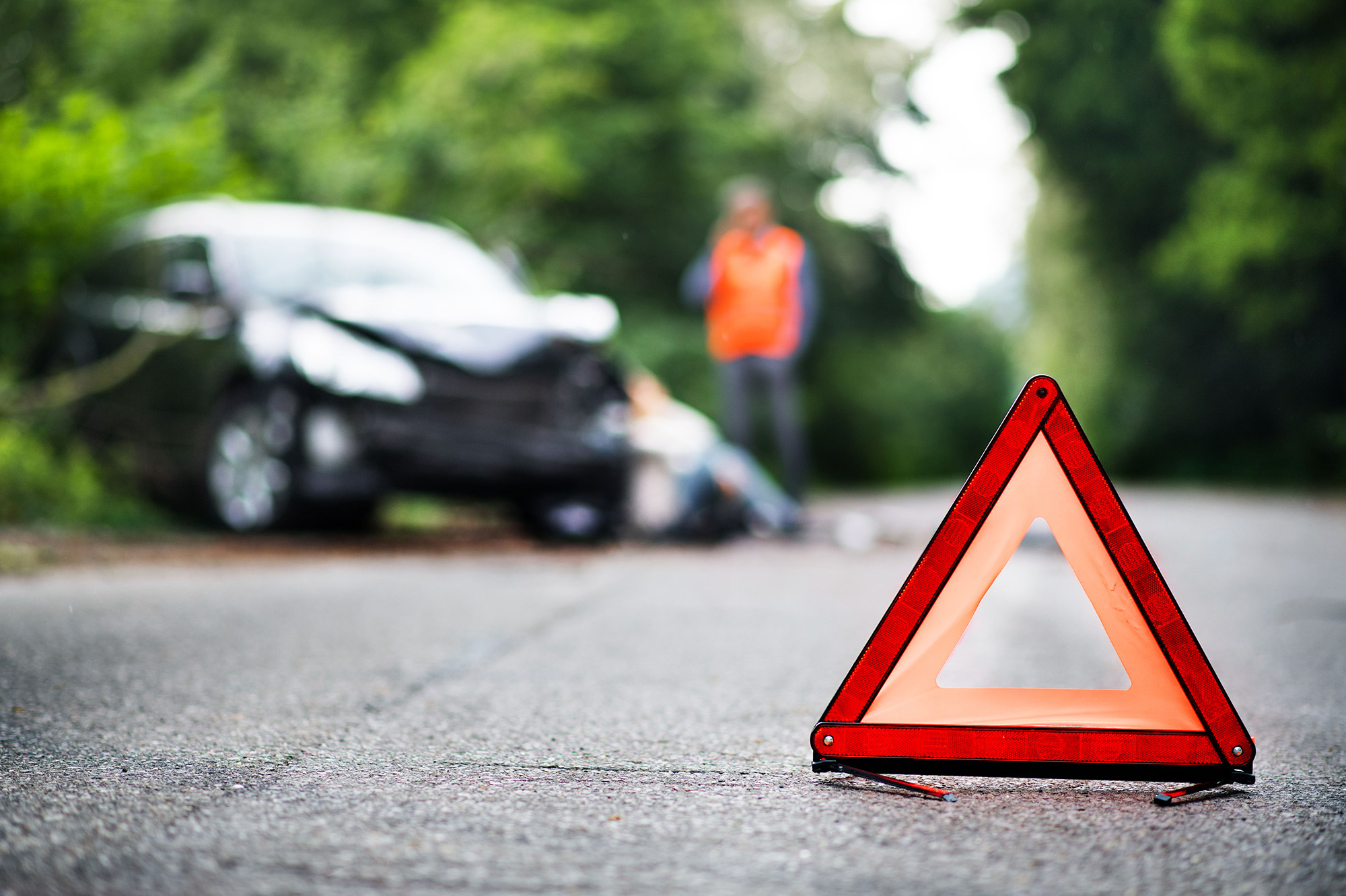 Red warning triangle on a road with a blurred damaged car and a person in an orange vest in the background.