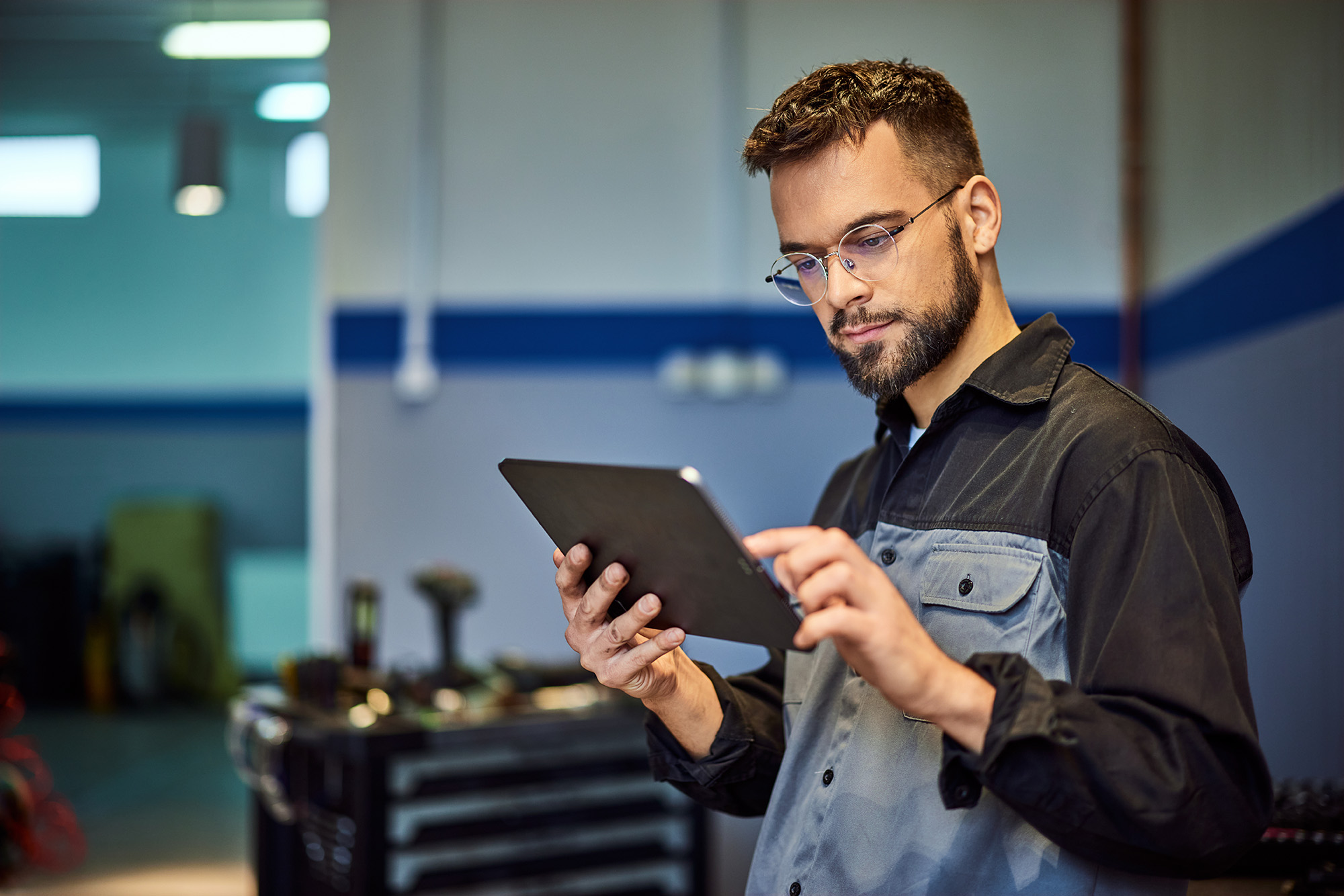 Man in a workshop wearing glasses and a work shirt using a digital tablet.