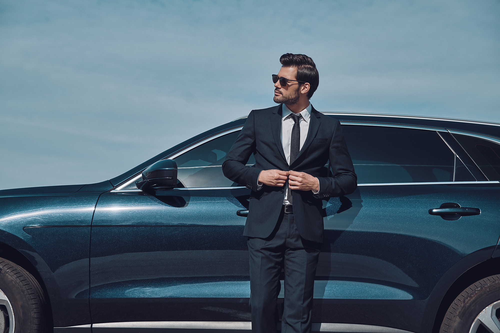 Man in a black suit and sunglasses standing against a dark blue luxury car under a clear sky.