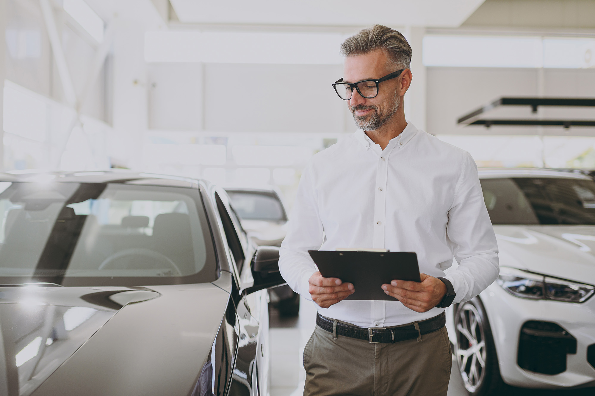Man in white shirt and glasses holding a clipboard standing next to a black car in a bright showroom.