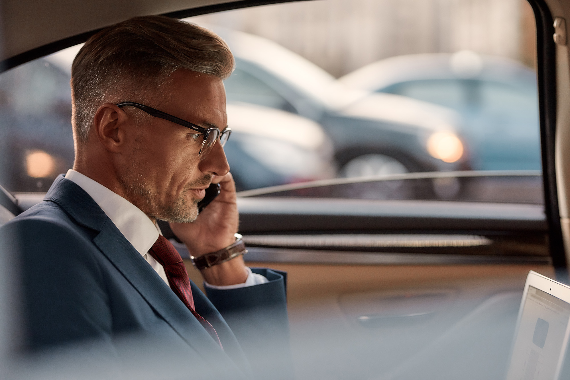 Middle-aged man in glasses and suit talking on a phone while looking at a laptop inside a car.