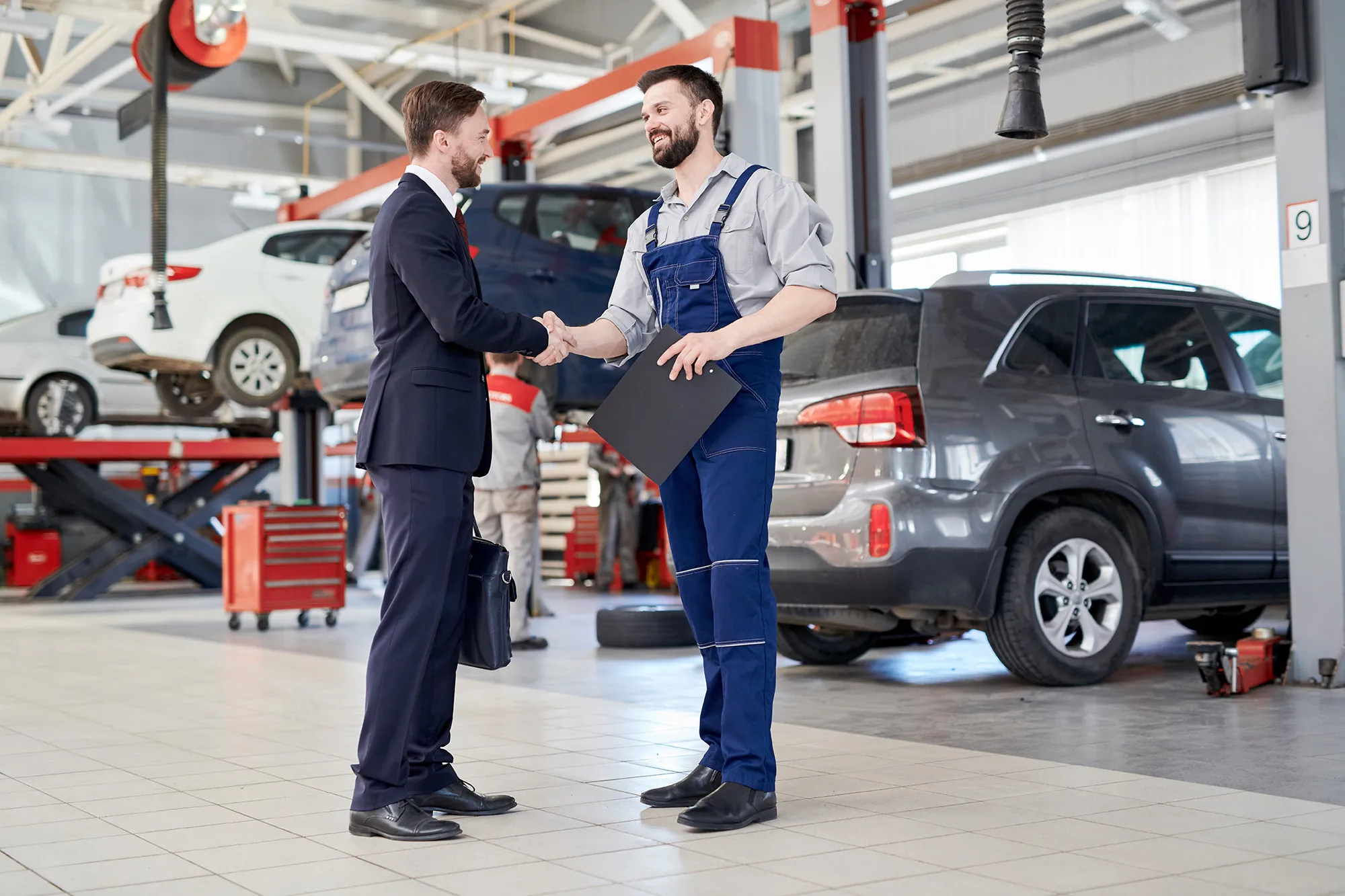 Man in business suit shaking hands with mechanic holding clipboard in a car repair shop.