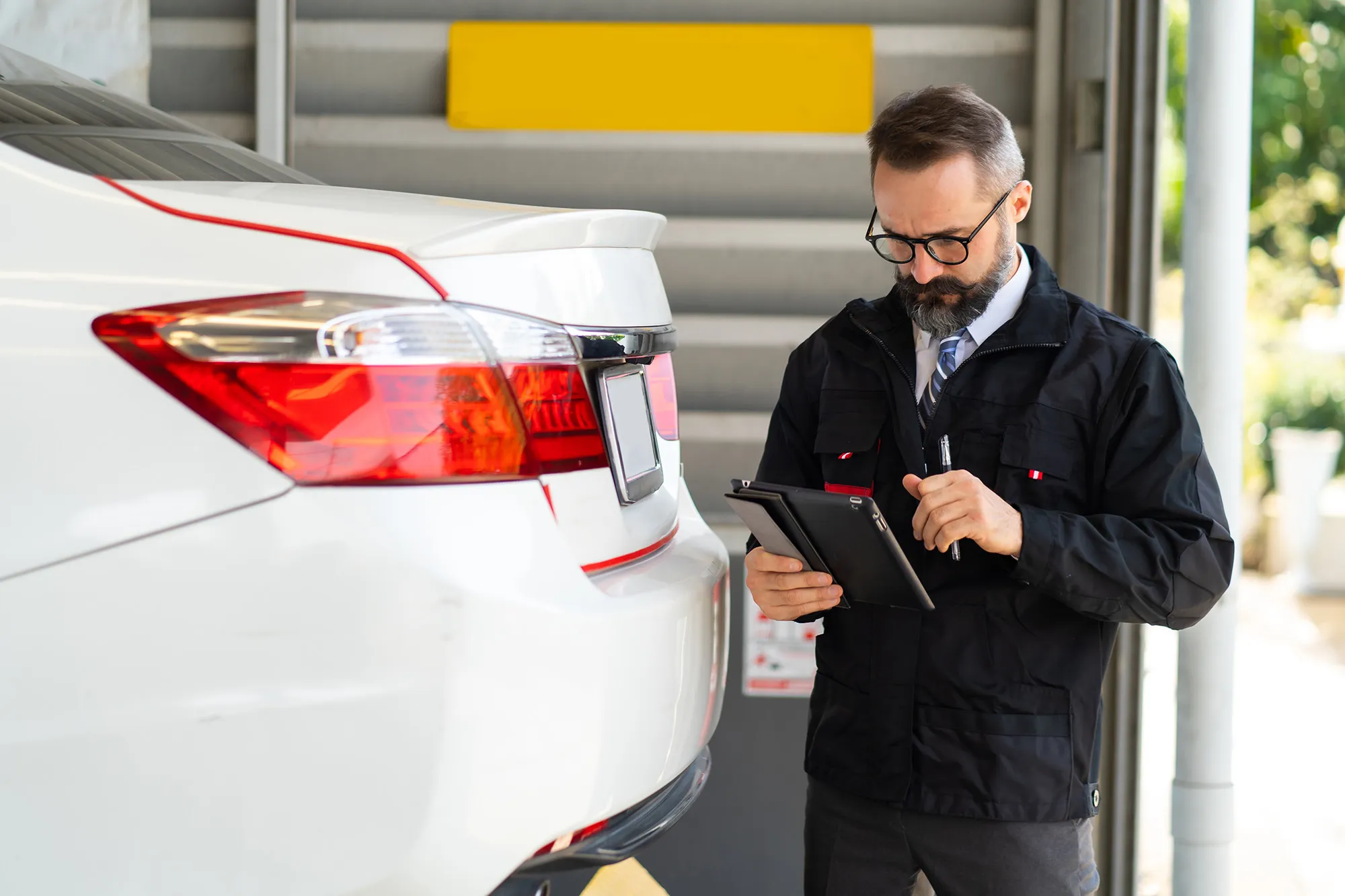 Man wearing glasses and a black jacket inspecting a white car's rear while using a tablet.