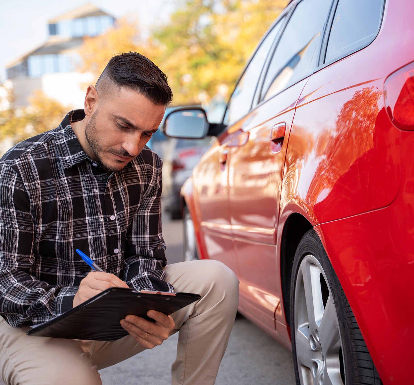 Man in a checkered shirt inspecting a red car and writing on a clipboard outdoors.