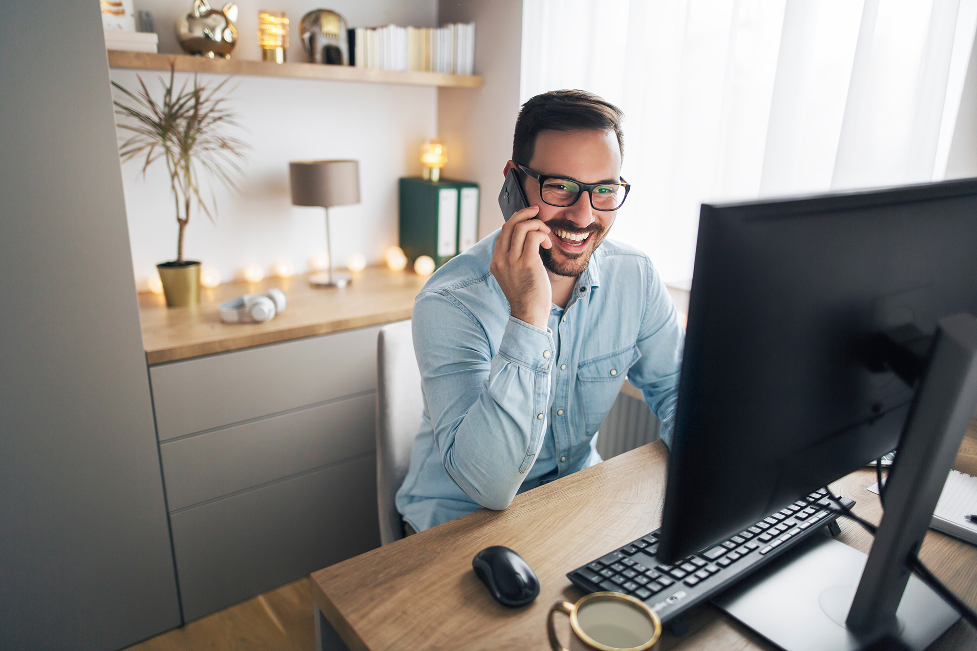 Man in glasses smiling and talking on phone while working at a desk with a computer in a bright office.