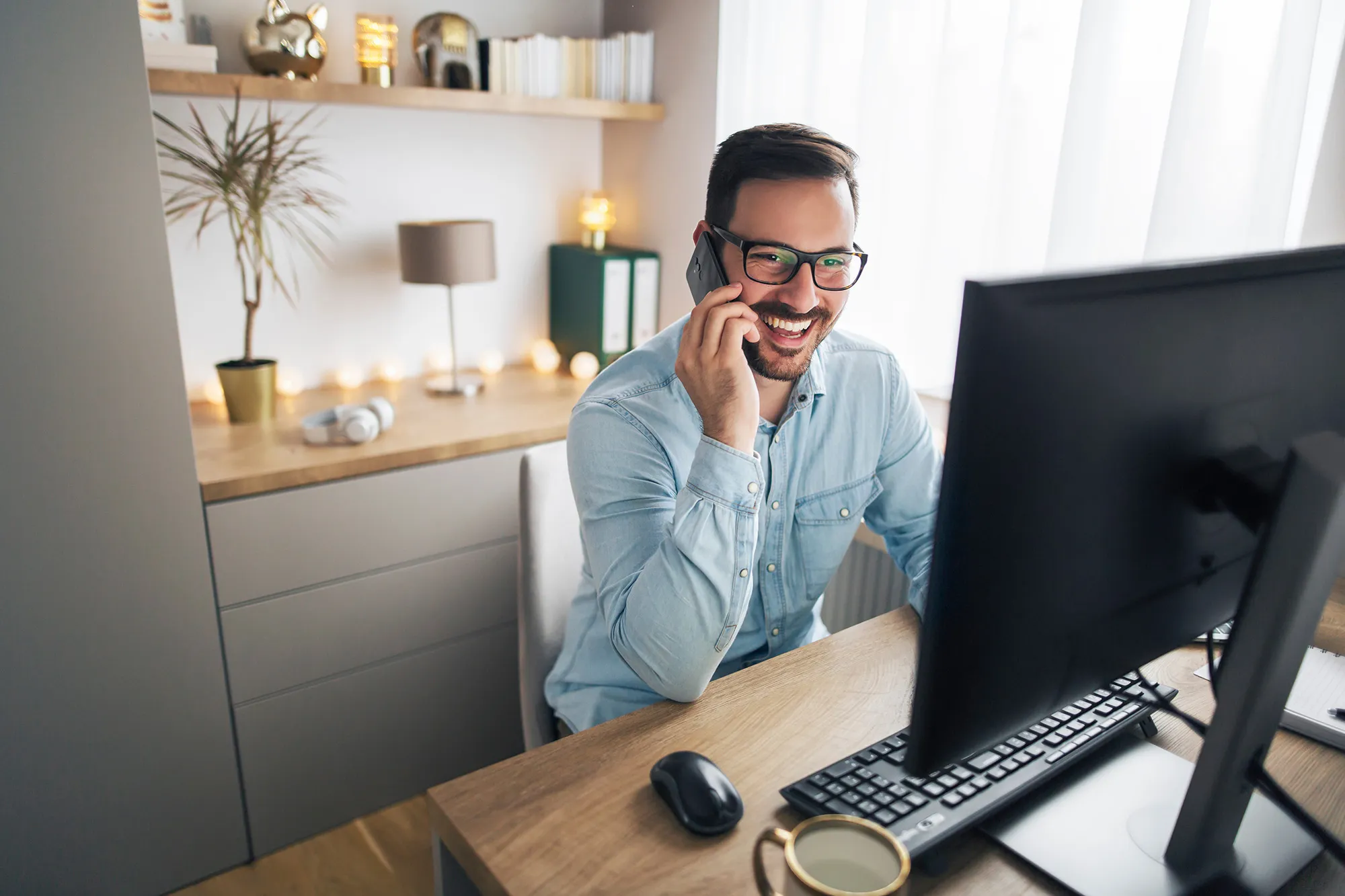 Man in glasses smiling and talking on phone while working at a desk with a computer in a bright office.