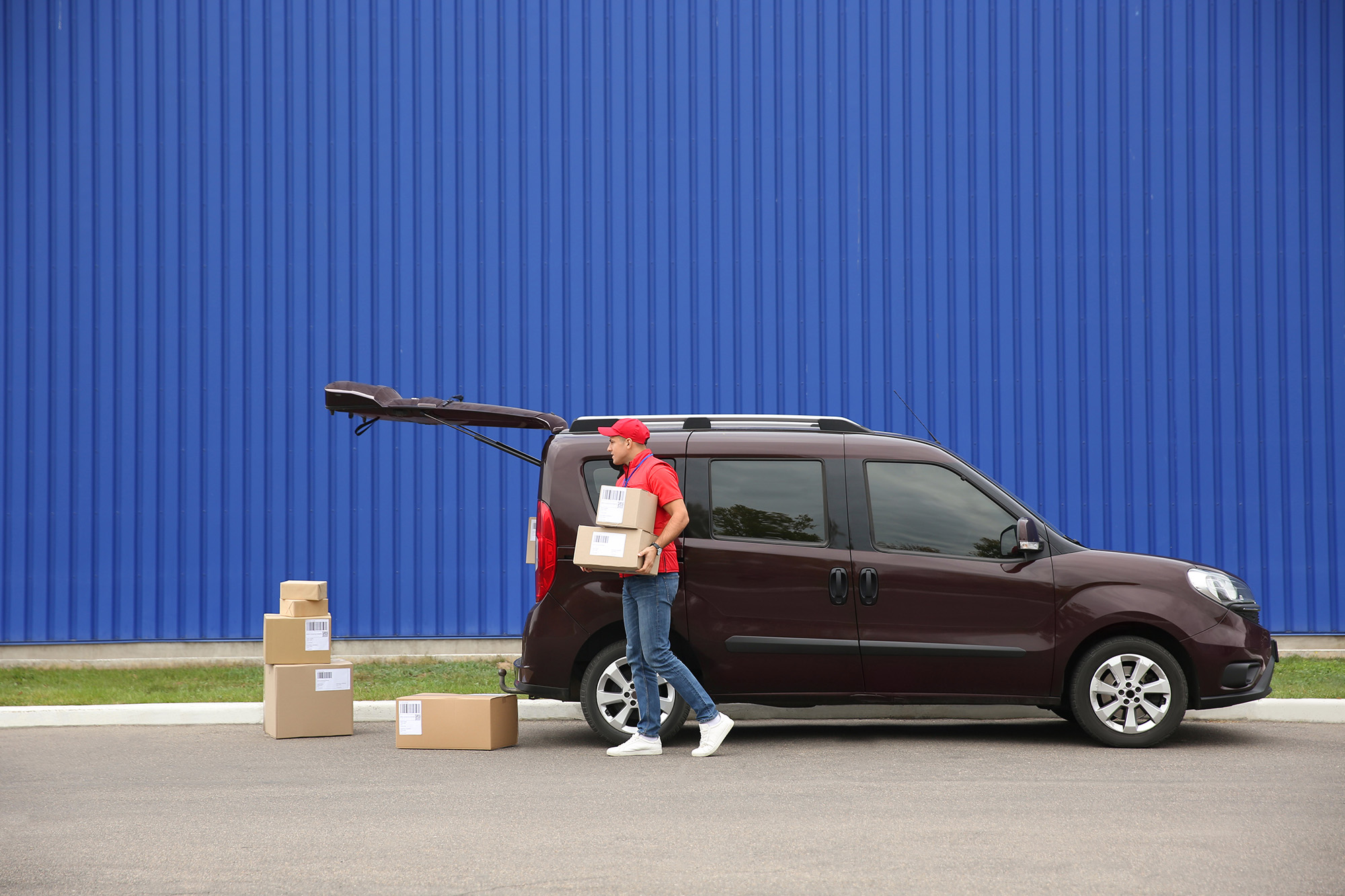 Delivery person in red uniform carrying two cardboard boxes from the open trunk of a maroon van parked in front of a blue corrugated wall.