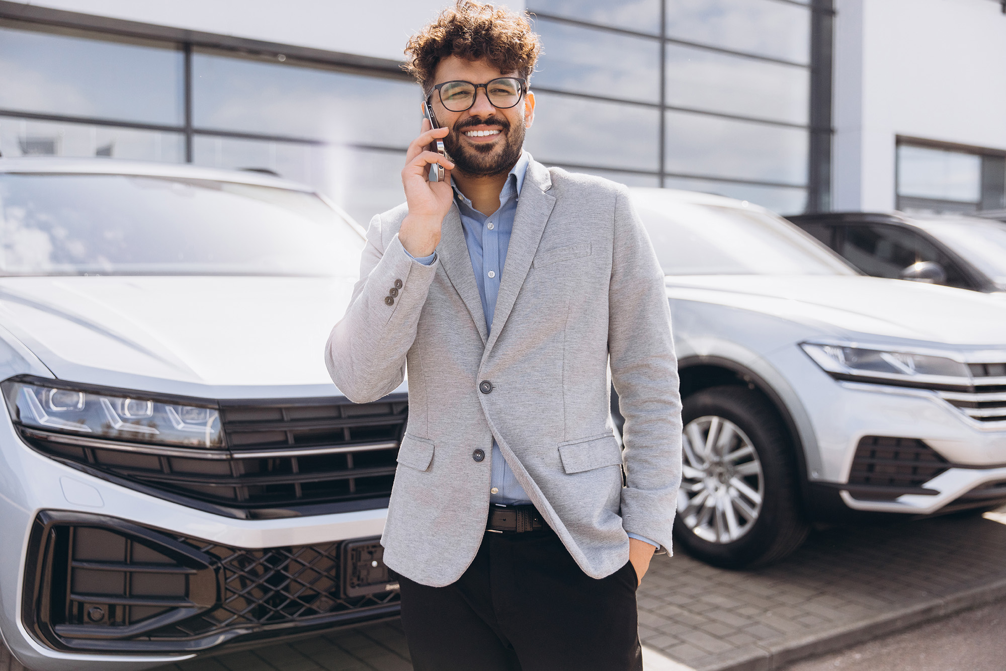 Smiling man in glasses and gray blazer talking on smartphone in front of parked modern silver cars.