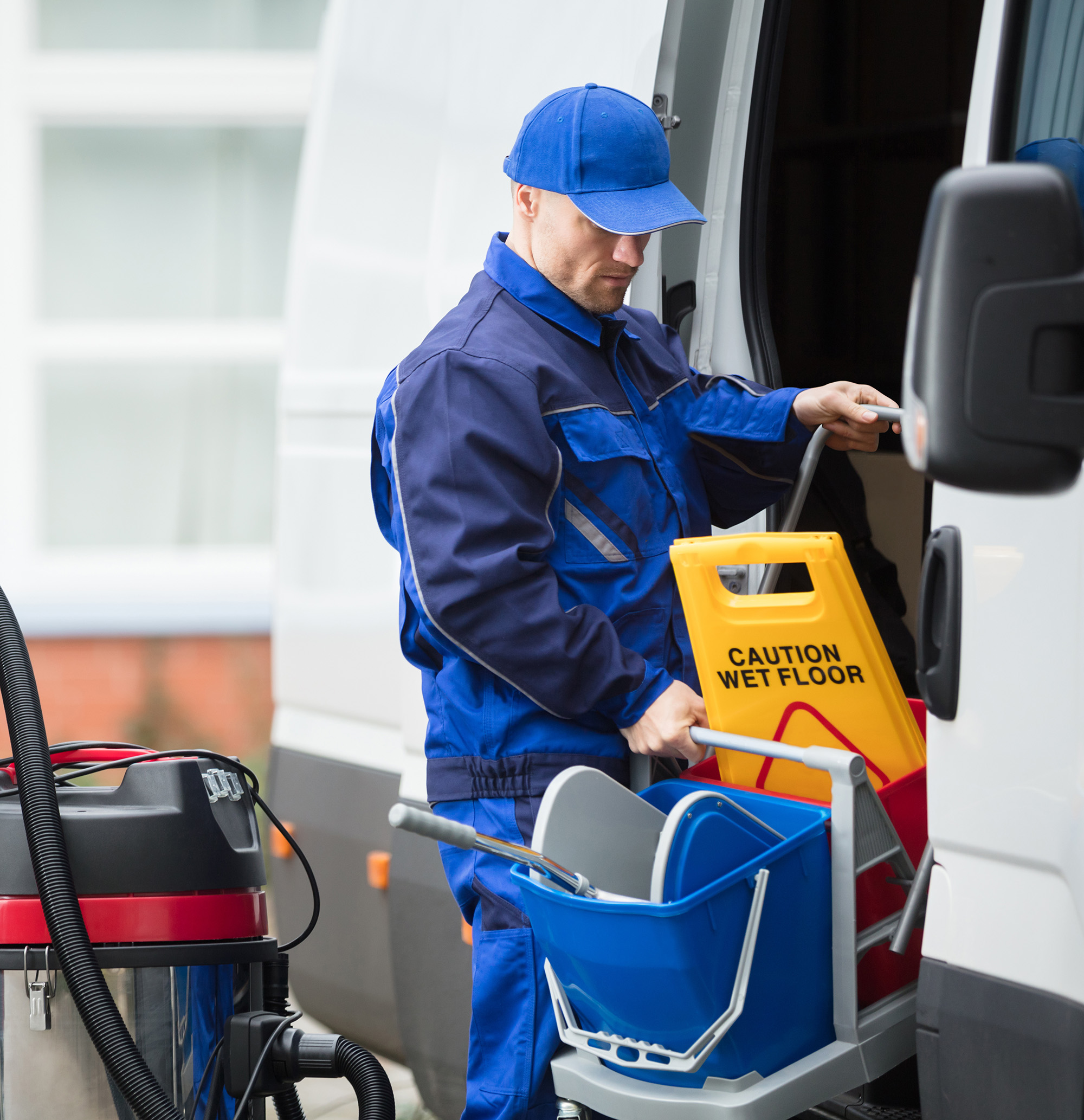 A worker in blue uniform loading cleaning supplies including a 'Caution Wet Floor' sign and mop bucket into a white van.