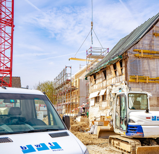 Construction site with a white construction van, excavator, scaffolding, and a crane lifting a platform near a partially renovated building.
