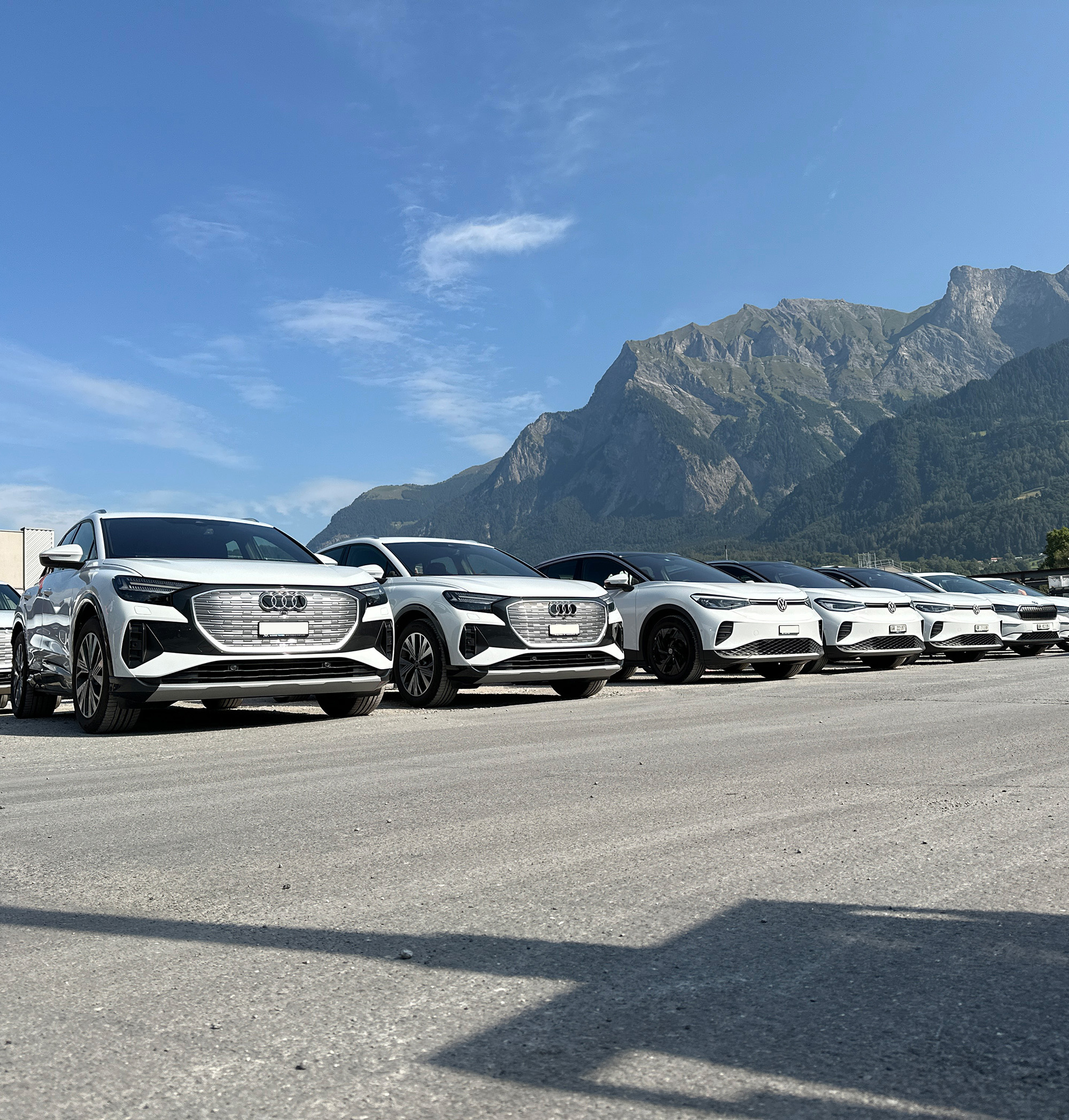 Row of white Audi and Volkswagen electric SUVs parked on asphalt with mountainous landscape under clear sky.
