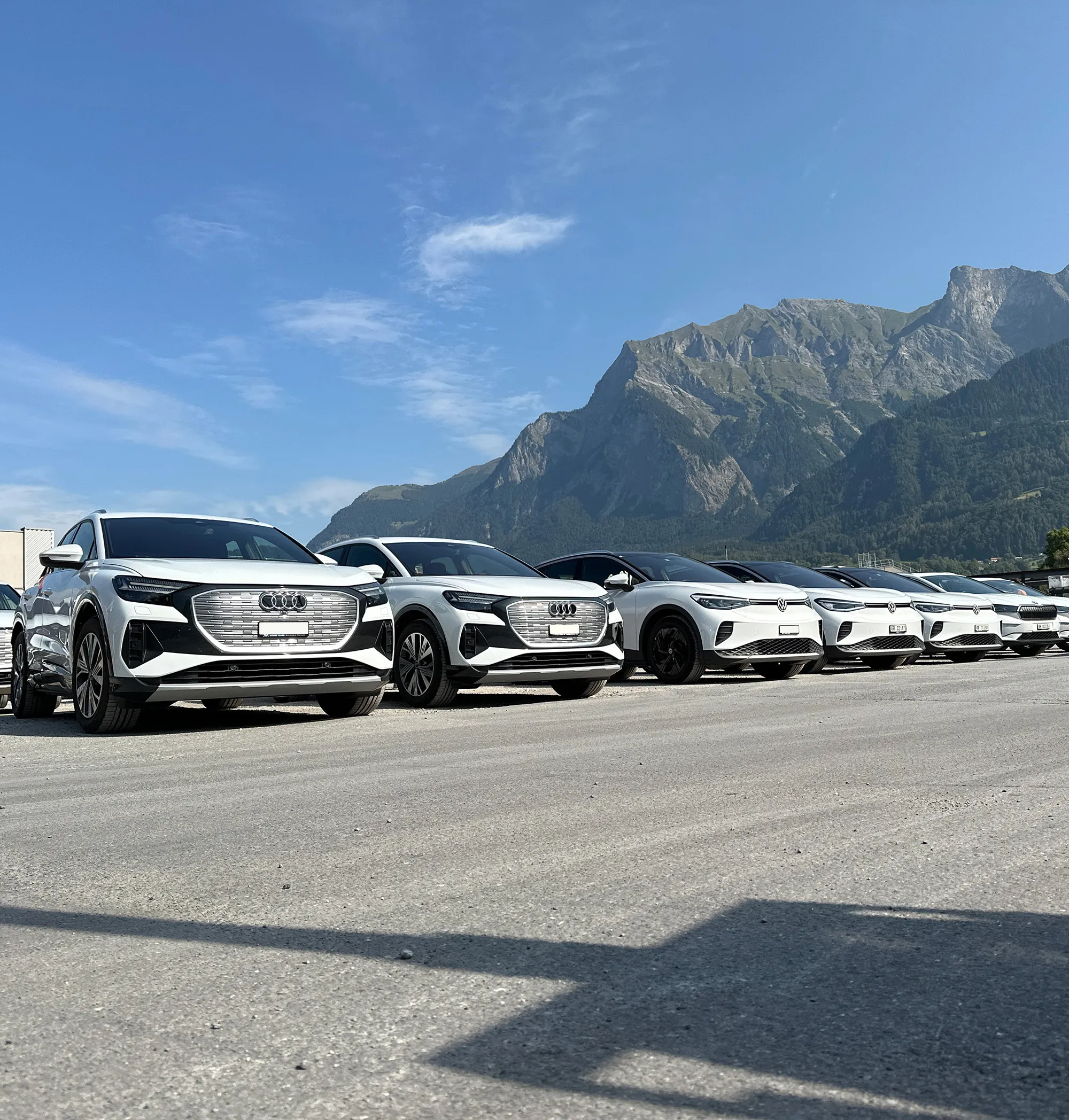 Row of white Audi and Volkswagen electric SUVs parked on asphalt with mountainous landscape under clear sky.