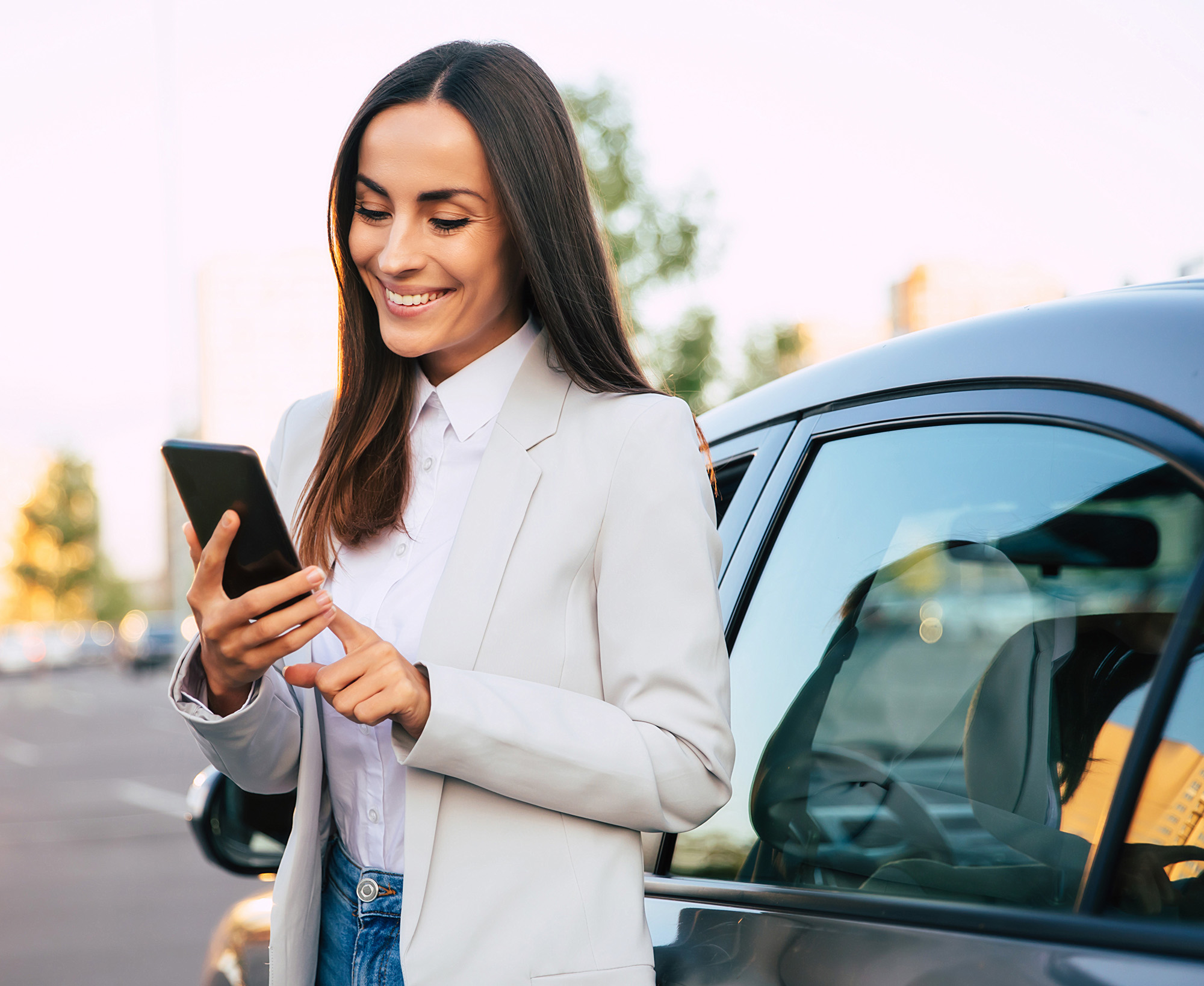 Smiling woman in a white blazer using a smartphone while standing next to a parked car.