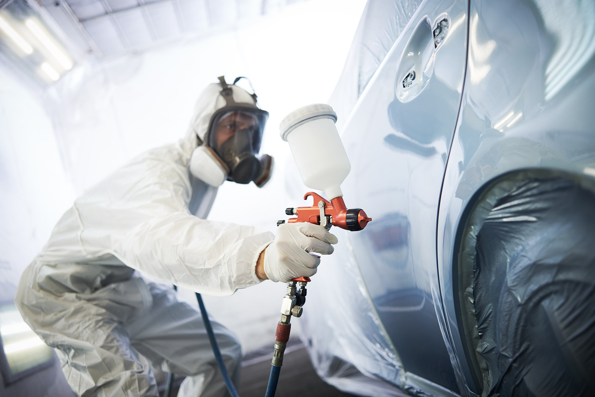 Person in protective suit and respirator spray painting a car door in a paint booth.