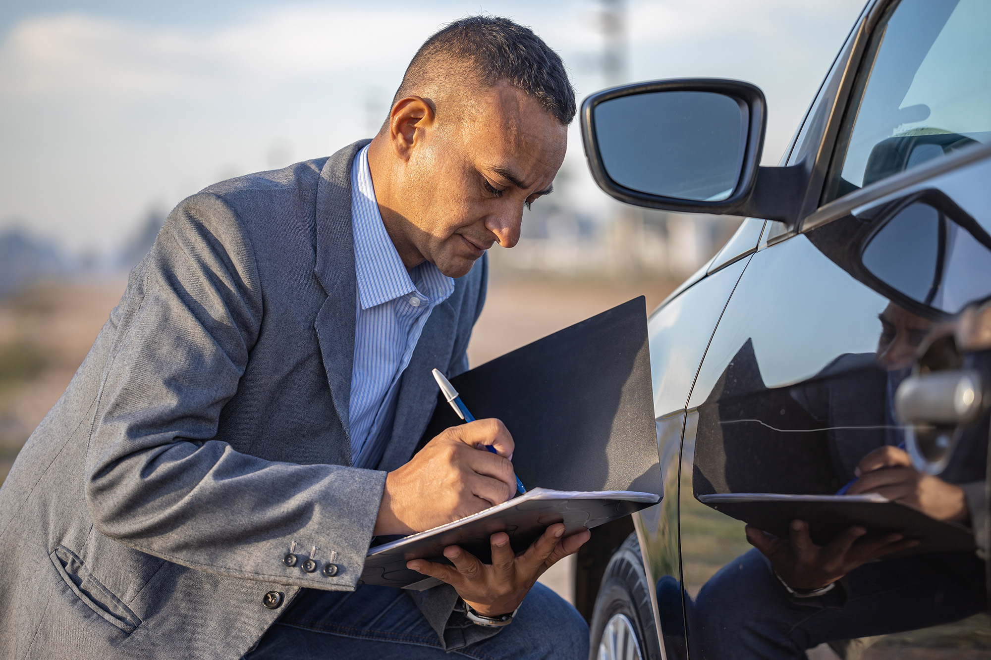 Man in a gray blazer inspecting a black car and writing notes on a clipboard.