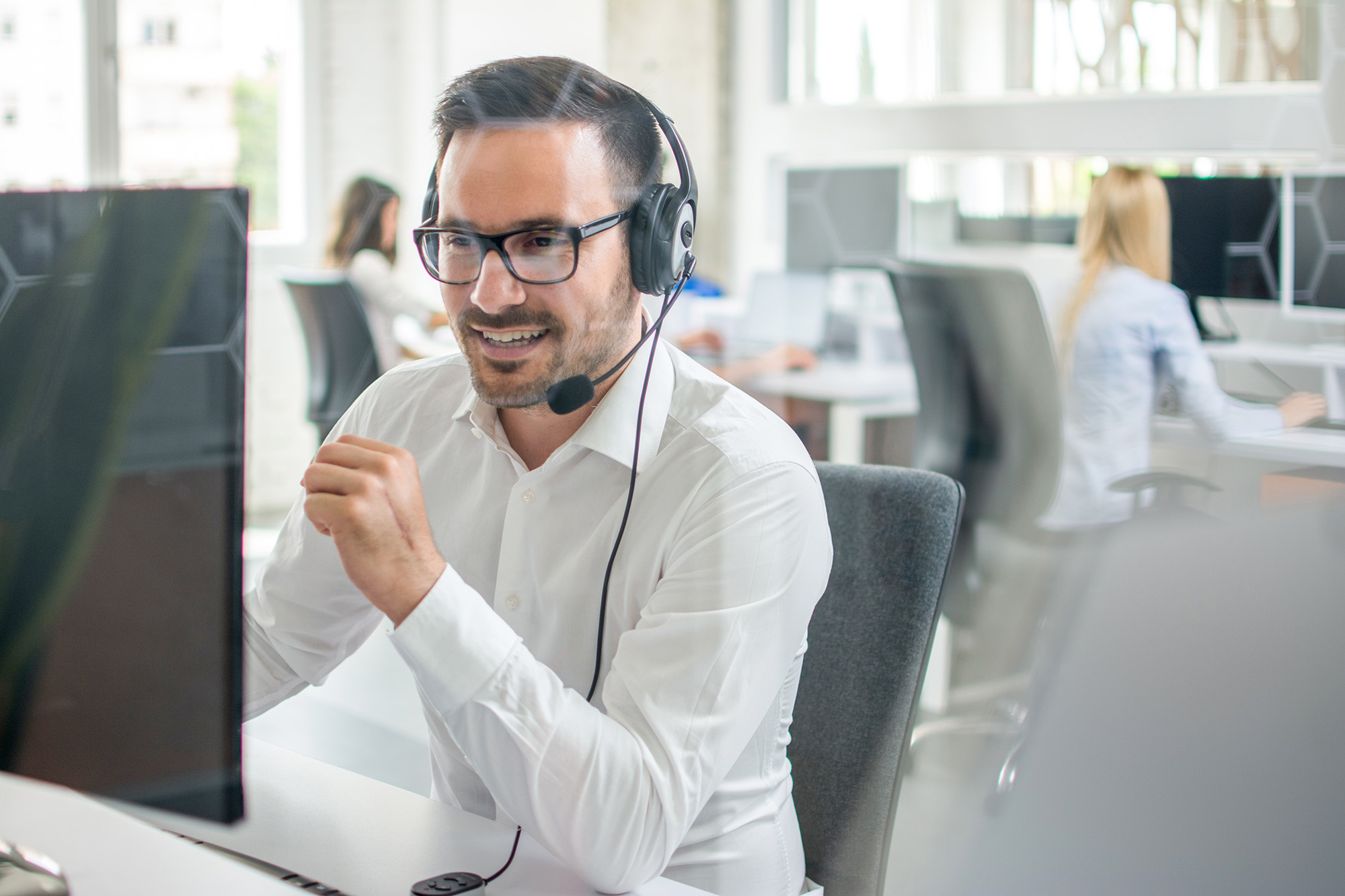 Man wearing glasses and headset, smiling and working at a computer in a modern office.