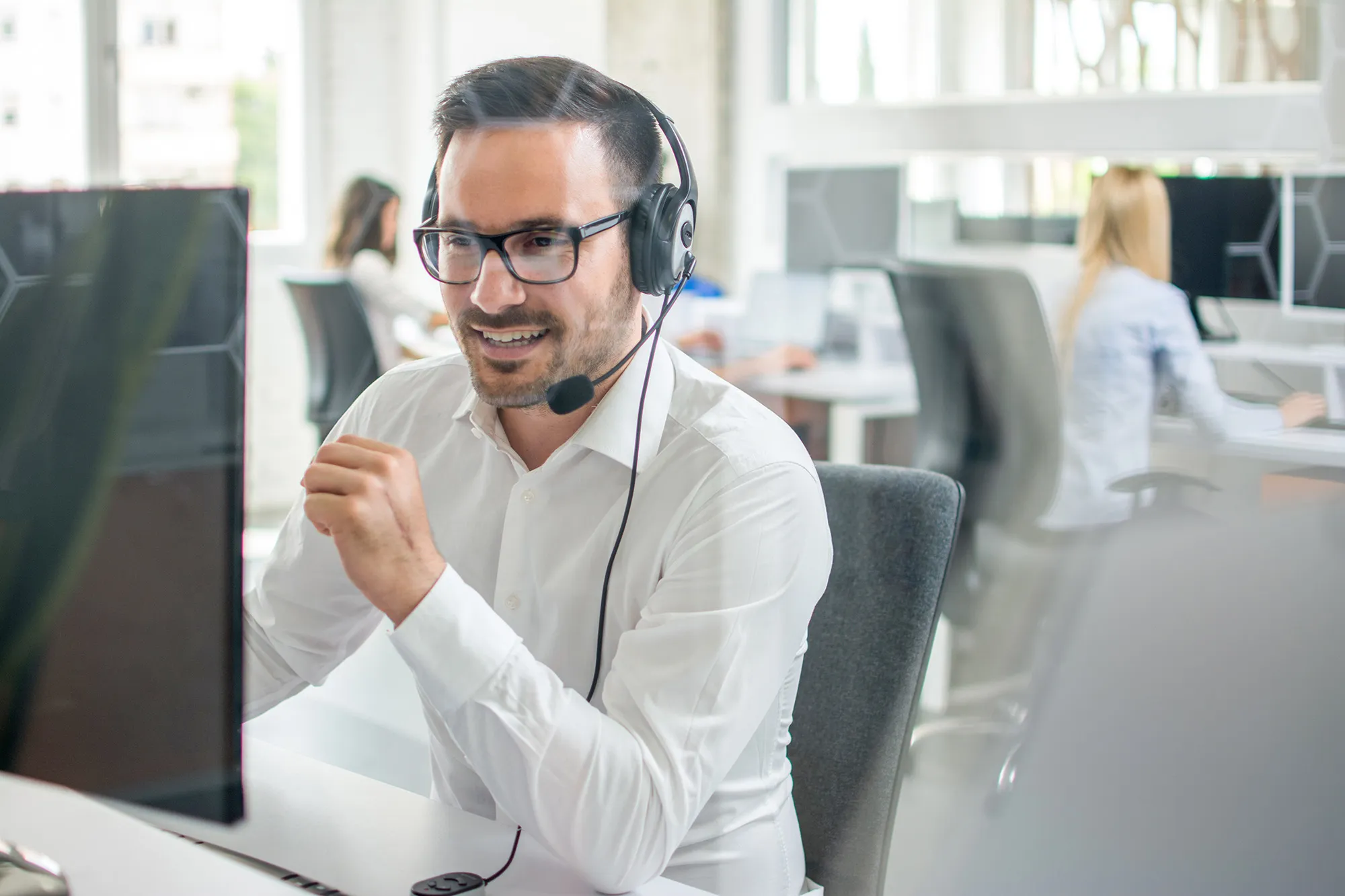 Man wearing glasses and headset, smiling and working at a computer in a modern office.