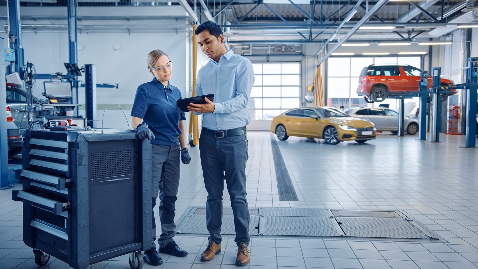 A male and female mechanic review information on a tablet next to a tool cart in a bright auto repair shop with cars in the background.