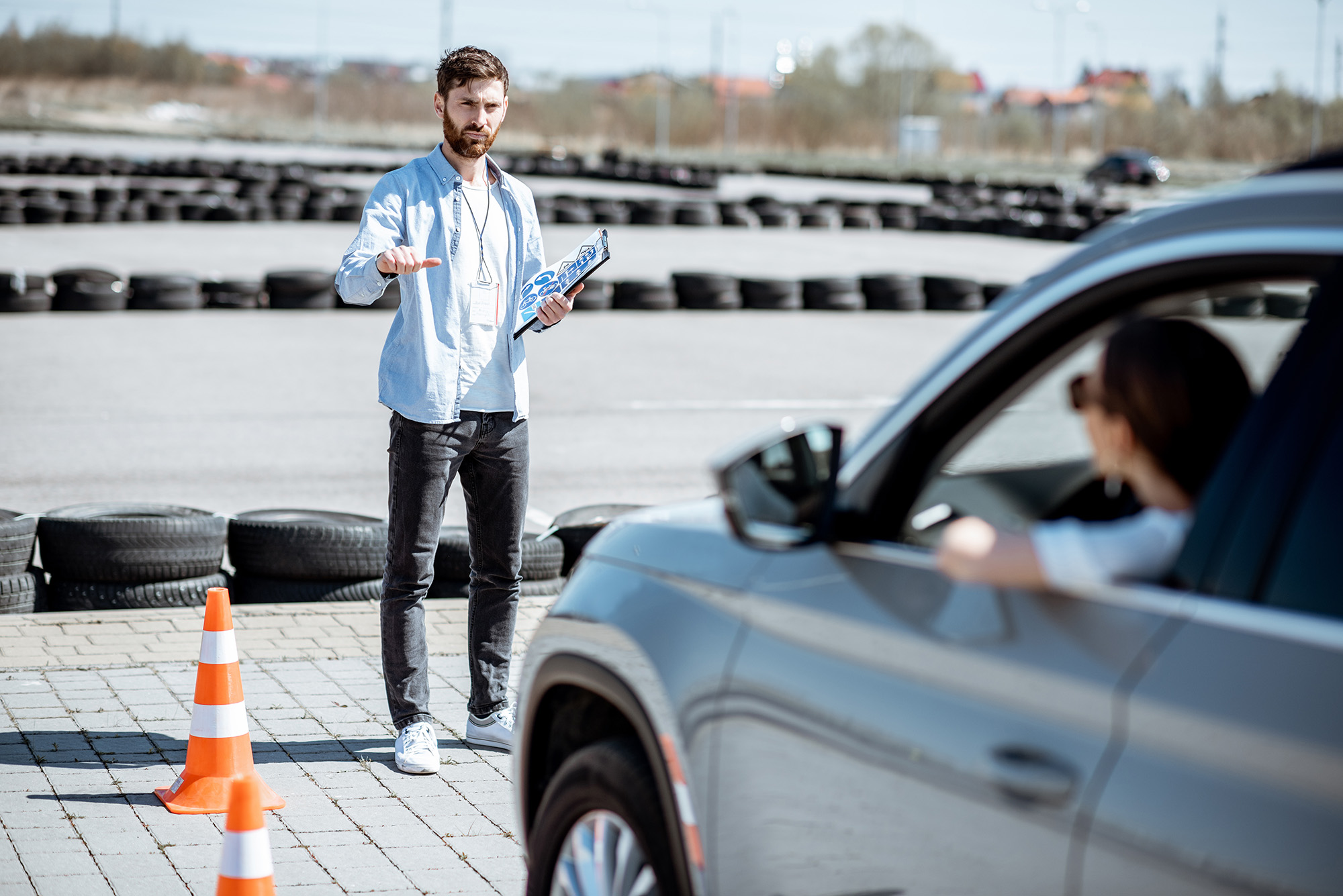 Man holding a clipboard and gesturing thumbs down at a driver in a gray SUV near orange traffic cones on a paved area with stacked tires in the background.