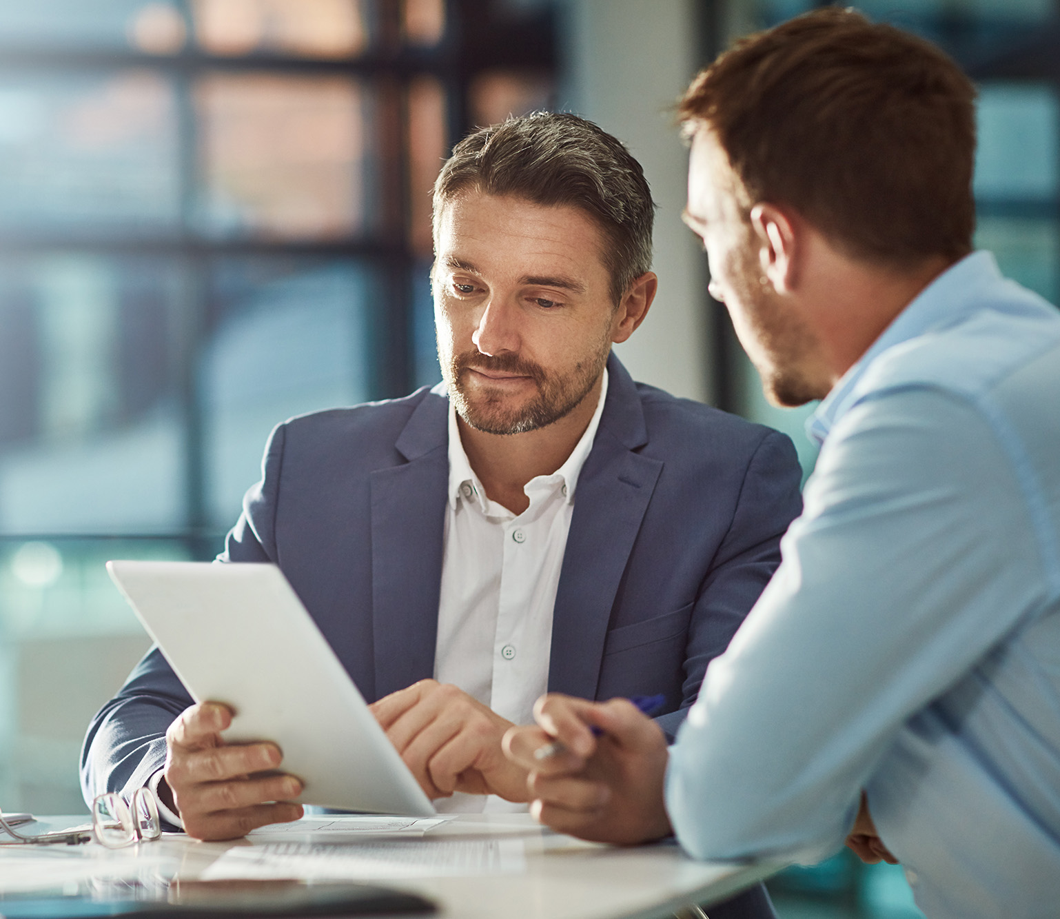 Two men in business attire reviewing a document on a tablet in an office setting.