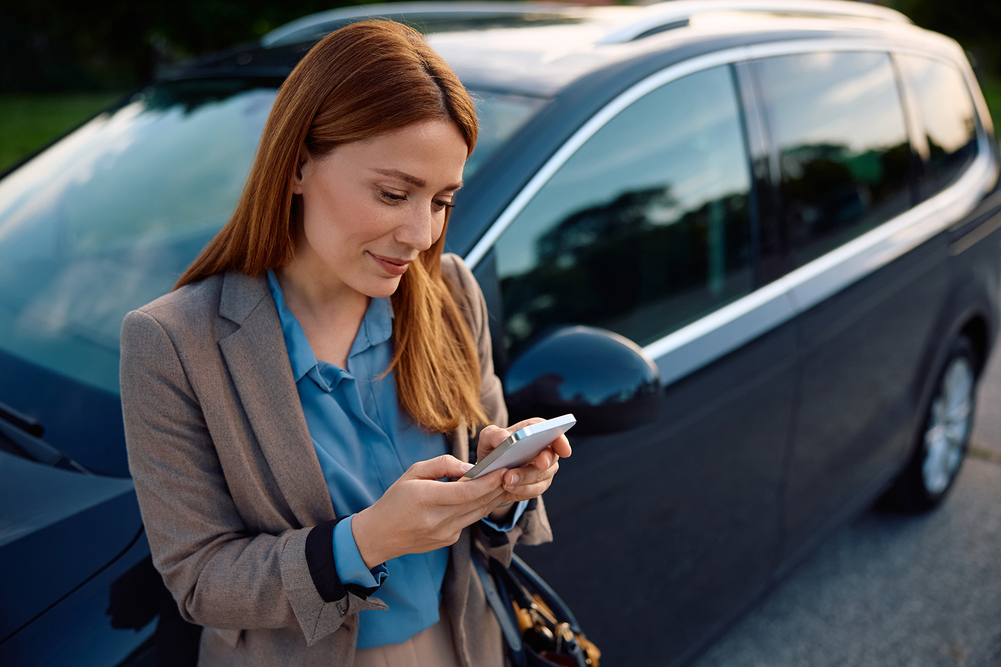 Woman in business attire using a smartphone standing next to a parked black car.