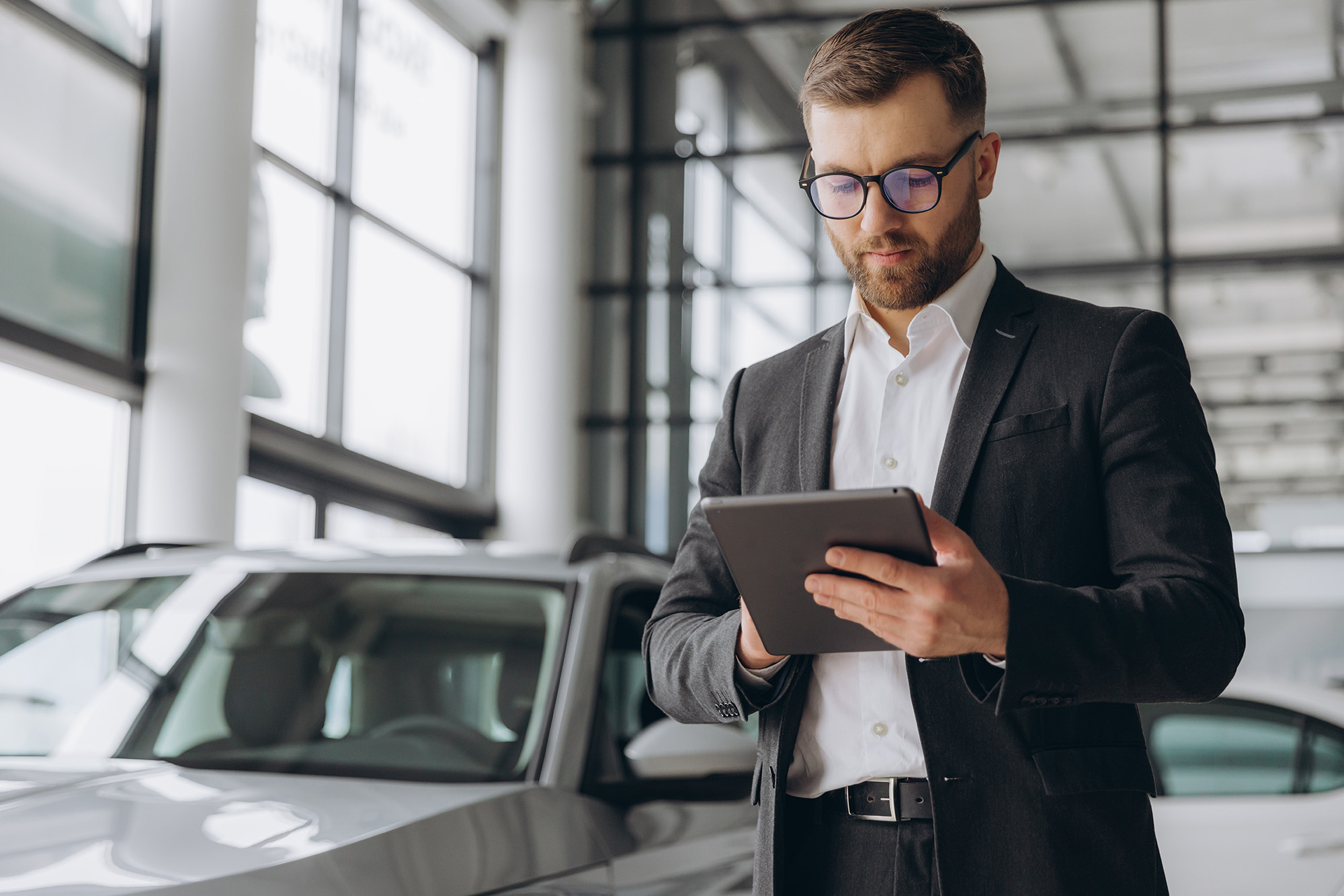 Man in glasses and business suit using a tablet while standing next to a silver car in a showroom.