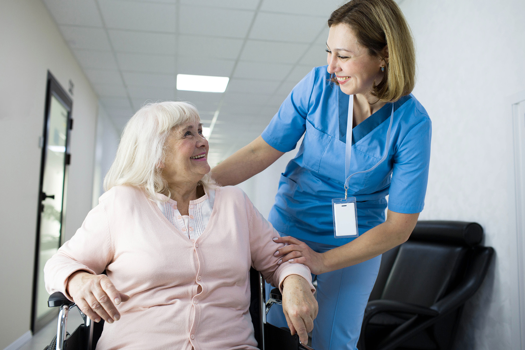 Smiling nurse in blue scrubs comforting and assisting an elderly woman in a wheelchair in a hallway.