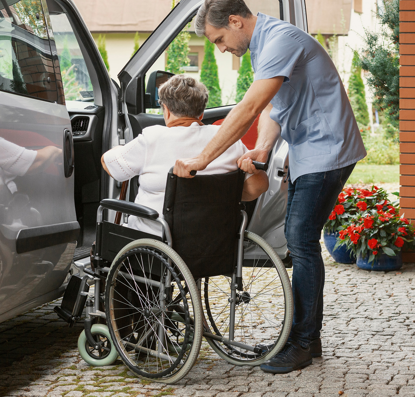 Caregiver assisting an elderly person in a wheelchair into a vehicle on a cobblestone driveway with flowers nearby.