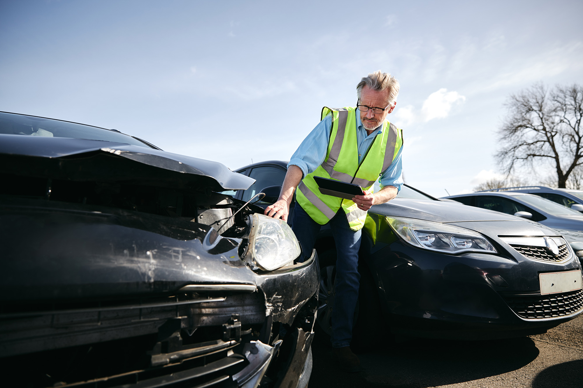 Man in a yellow safety vest inspecting the front damage of a black car while holding a tablet outside on a sunny day.