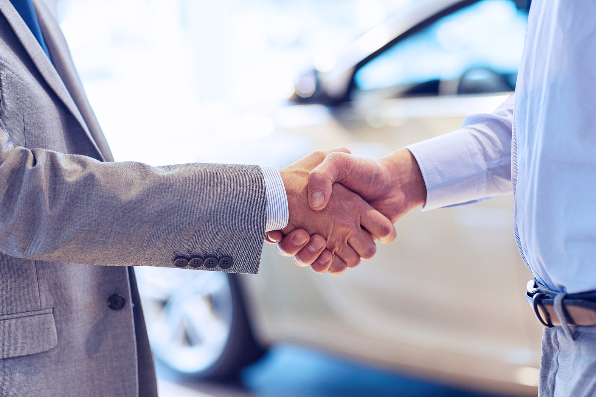 Two people shaking hands in front of a car, one person wearing a suit and the other wearing a light blue shirt.