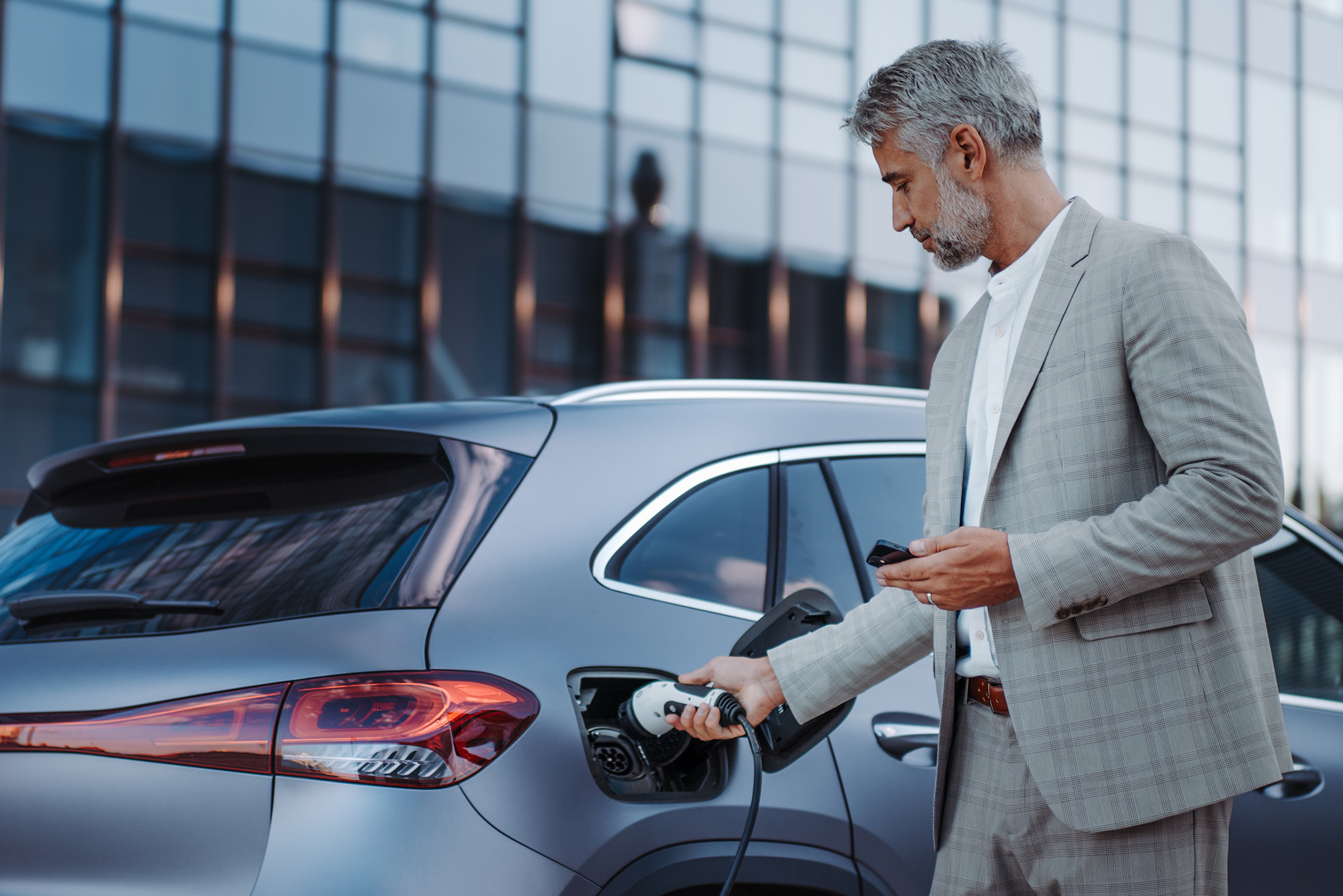 Man in a gray suit plugging a charging cable into an electric car outside a modern building.