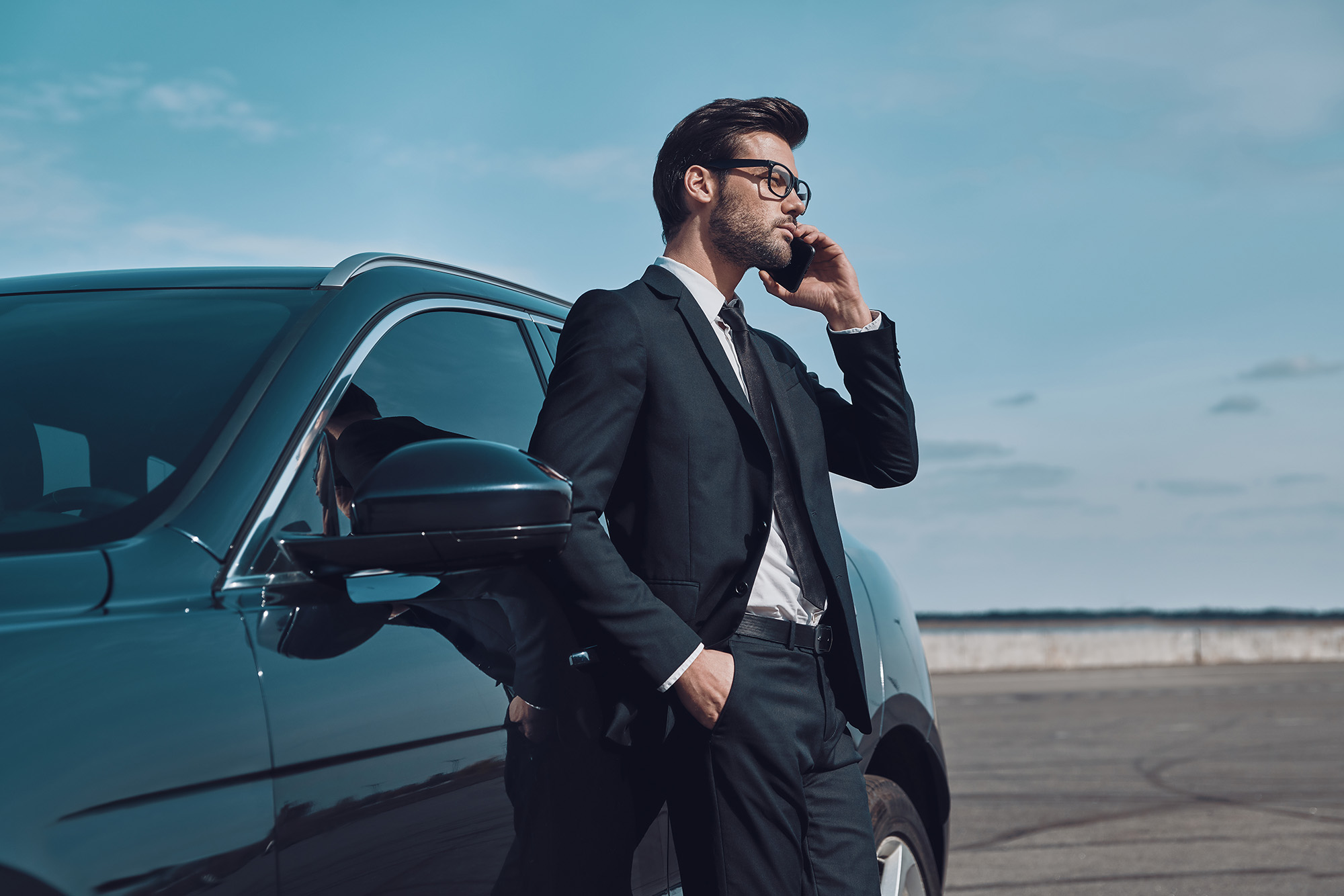 Man in black suit and glasses standing next to a black car, talking on a mobile phone outdoors.