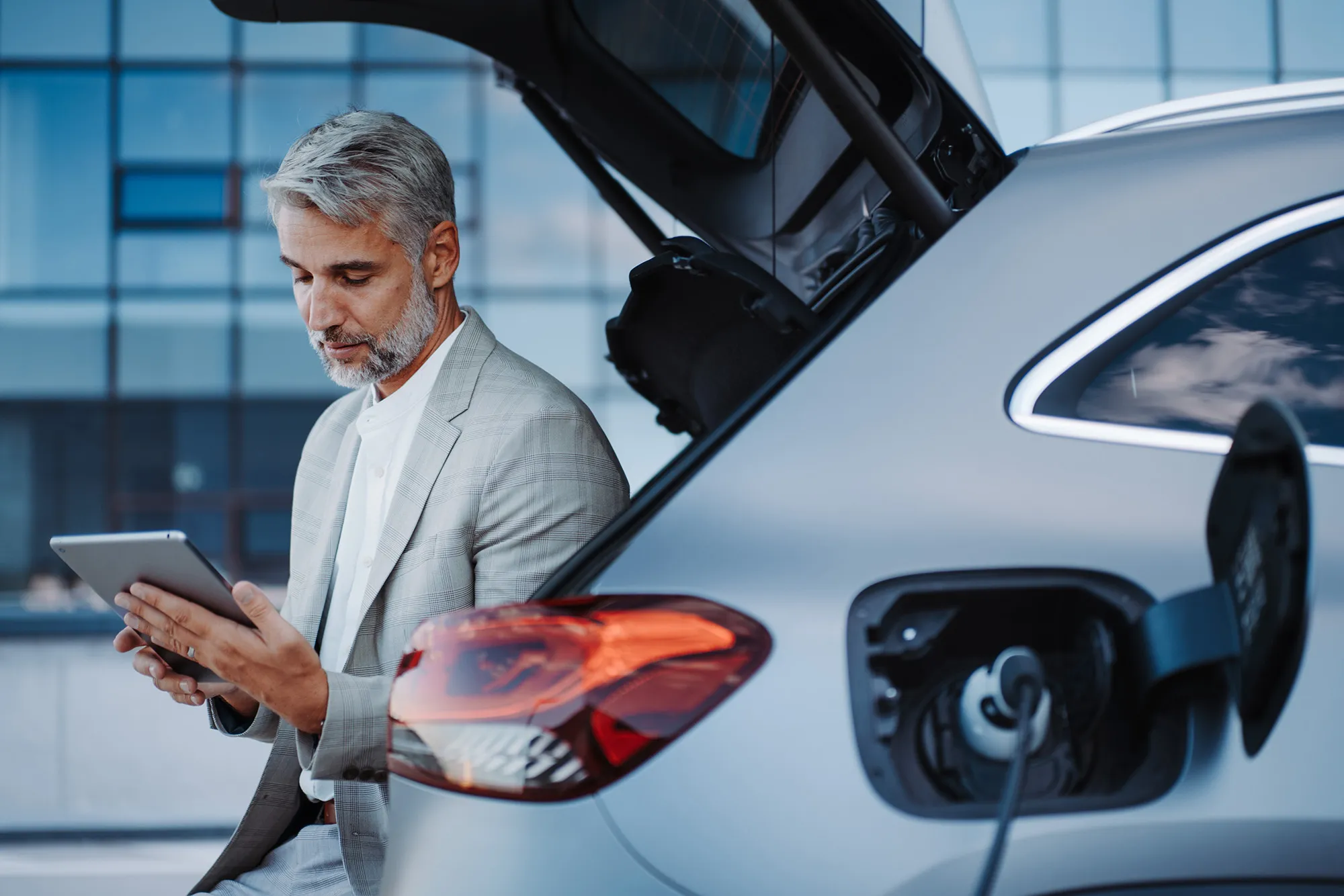 Man in gray suit using a tablet next to a silver electric car charging with the trunk open.