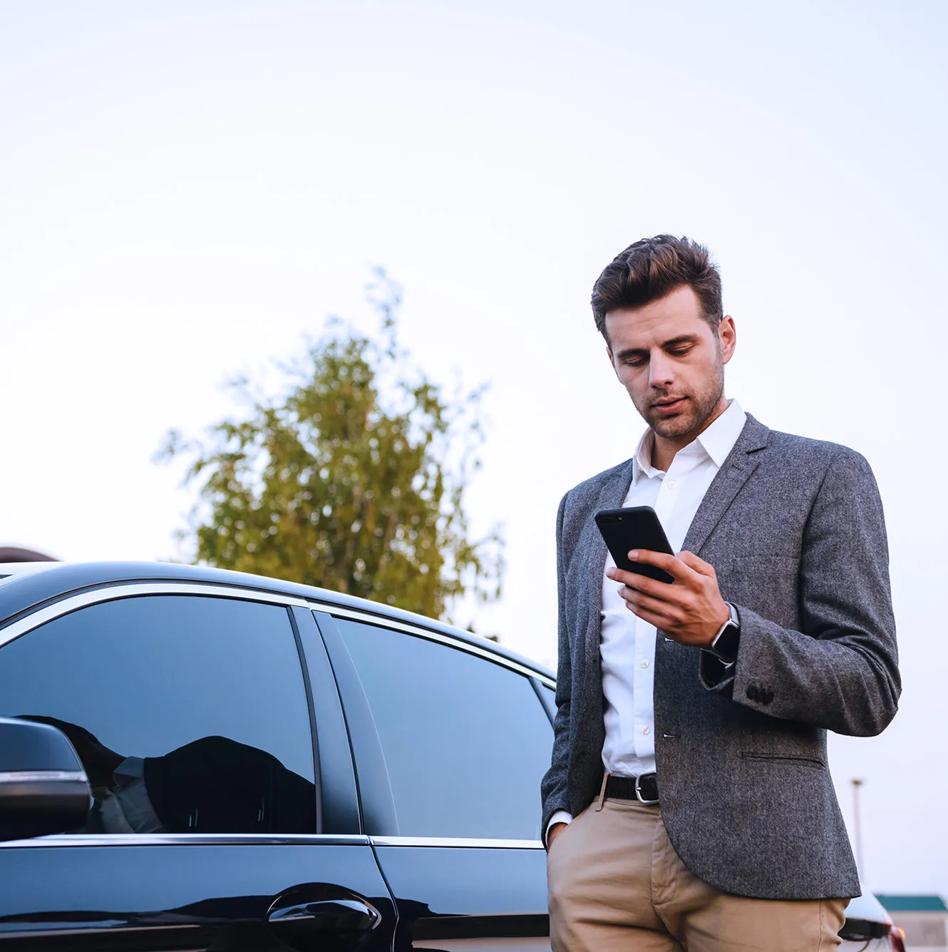 Man in a blazer and beige pants standing beside a black car, looking at his smartphone.