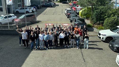 Group of about 35 people standing outdoors on pavement in front of parked cars, waving and smiling at the camera.