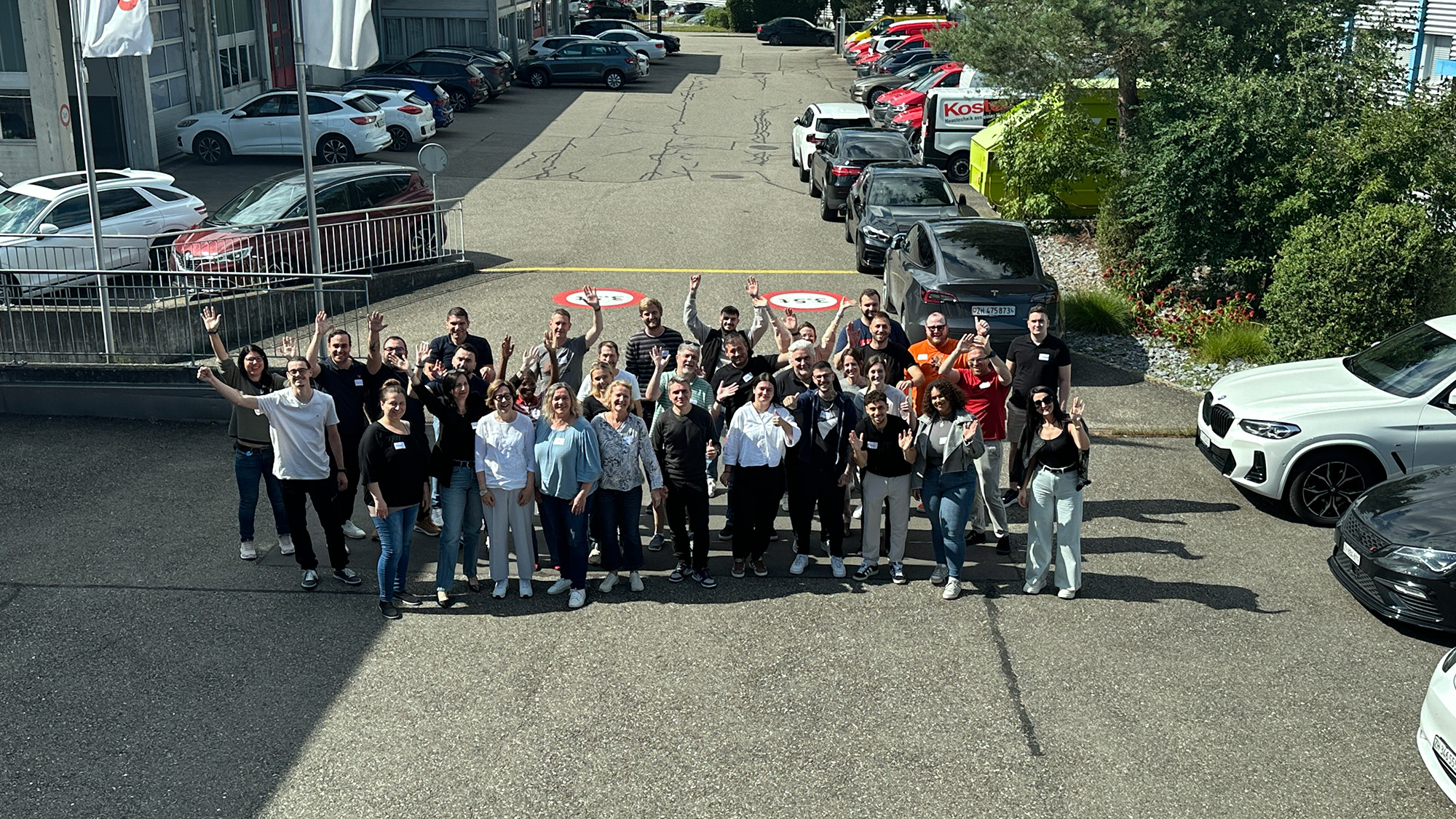 Group of about 35 people standing outdoors on pavement in front of parked cars, waving and smiling at the camera.