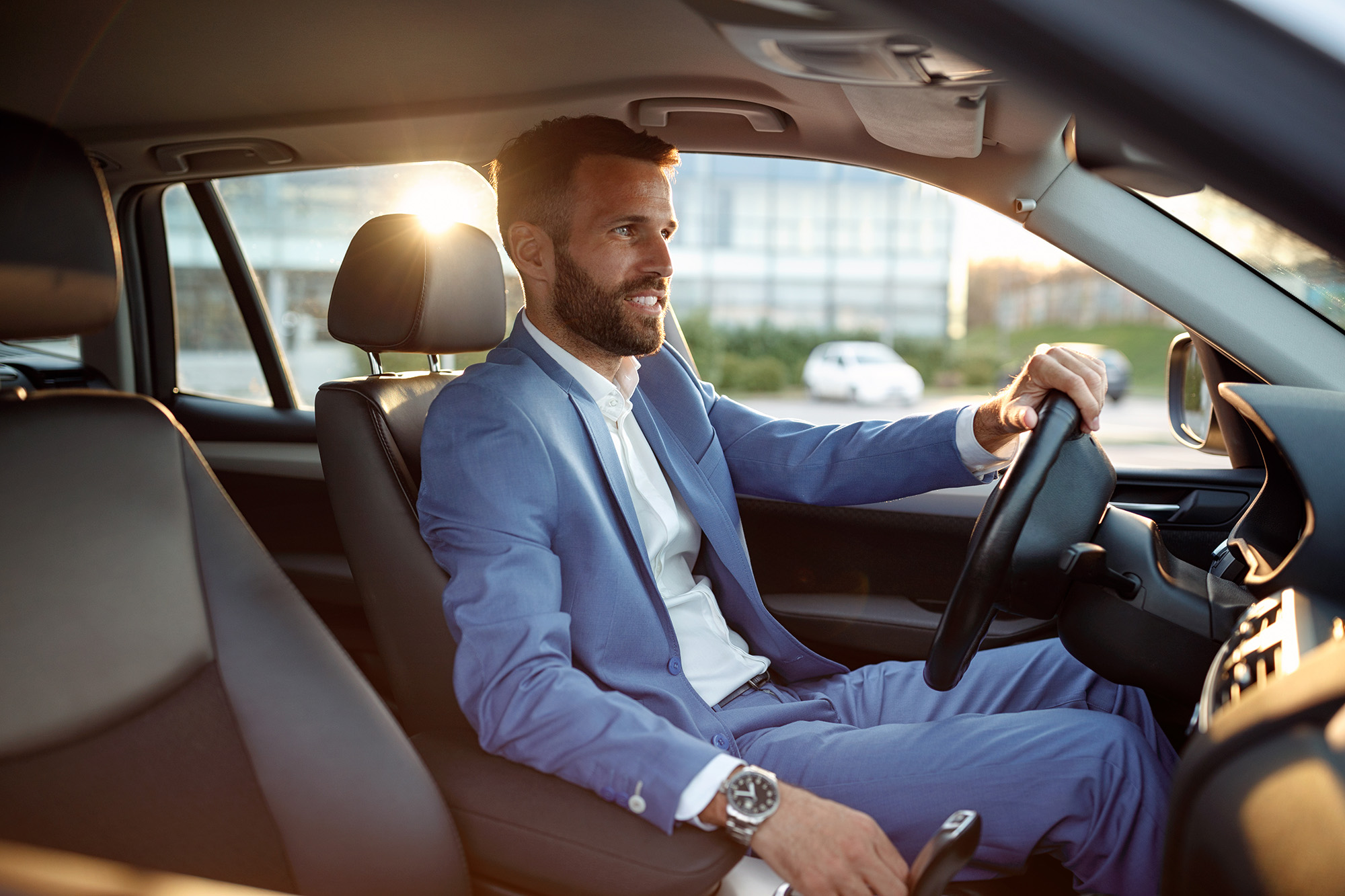 A man in a blue suit driving a car with sunlight coming through the window behind him.
