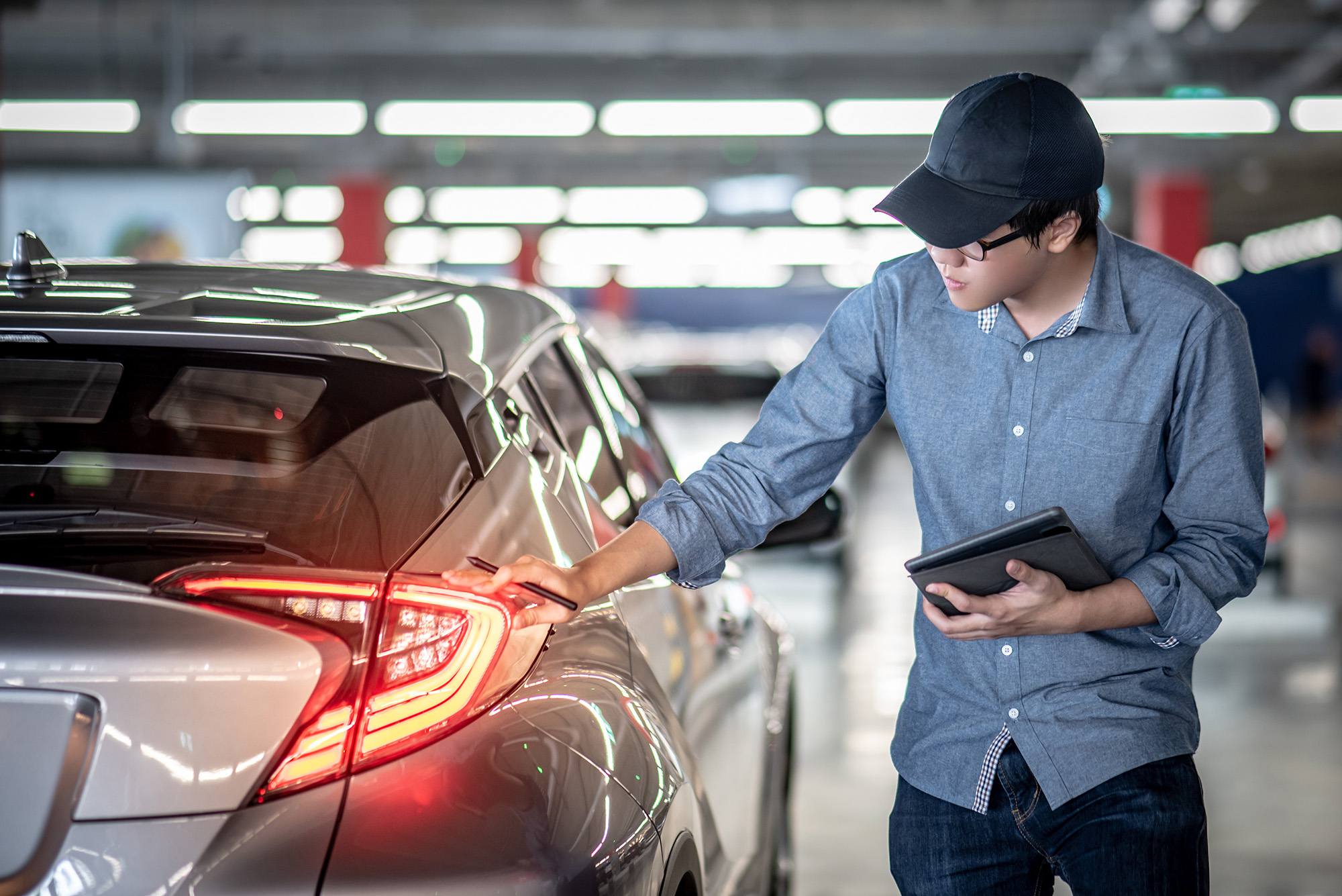Man in a blue shirt and black cap inspecting a car's rear light in a parking garage while holding a tablet.