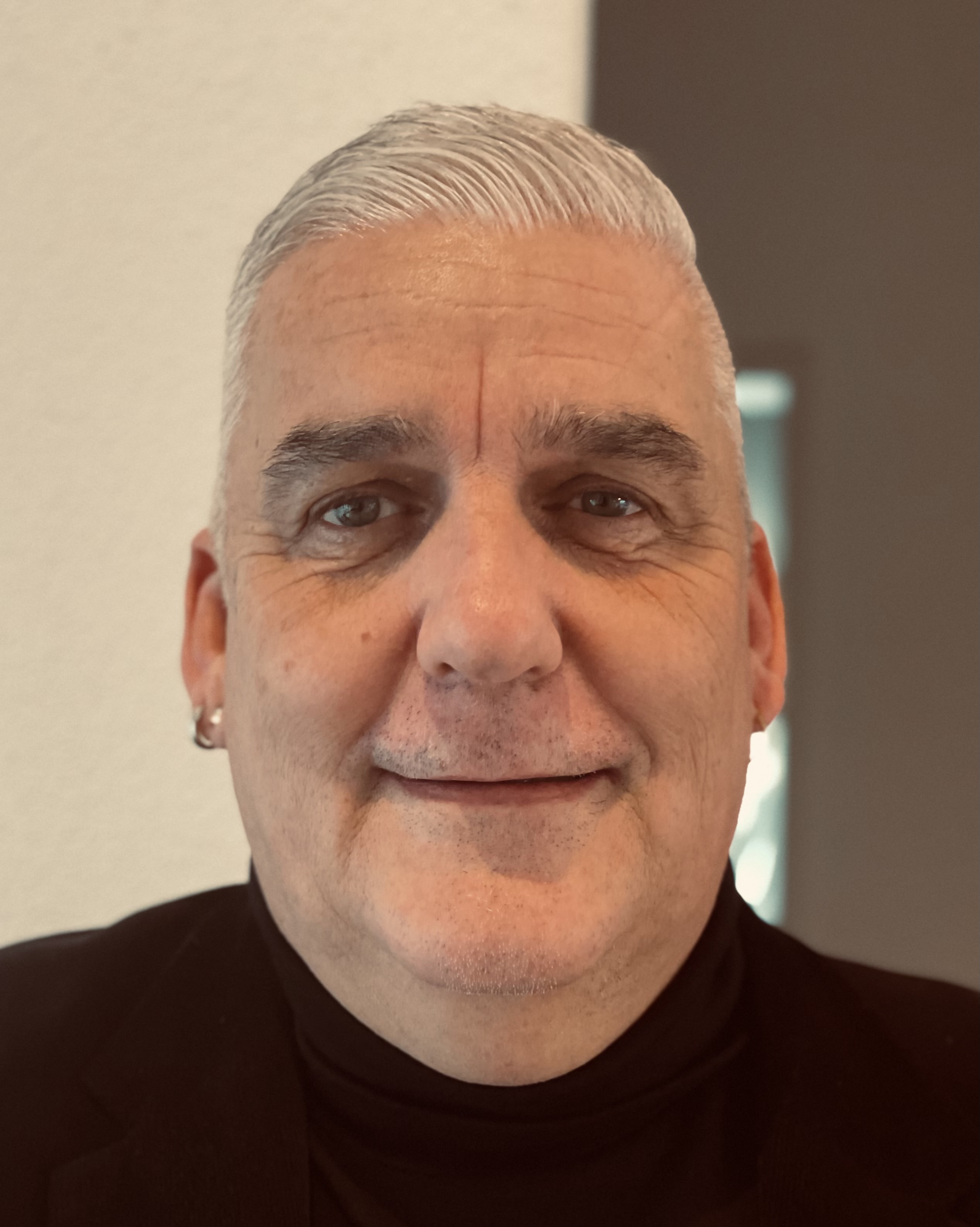 Close-up portrait of a middle-aged man with short gray hair, wearing a black turtleneck and blazer, smiling slightly.