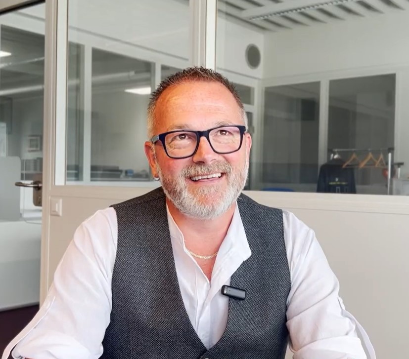 Smiling middle-aged man with glasses, gray beard, and short hair wearing a white shirt and gray vest in an office setting.