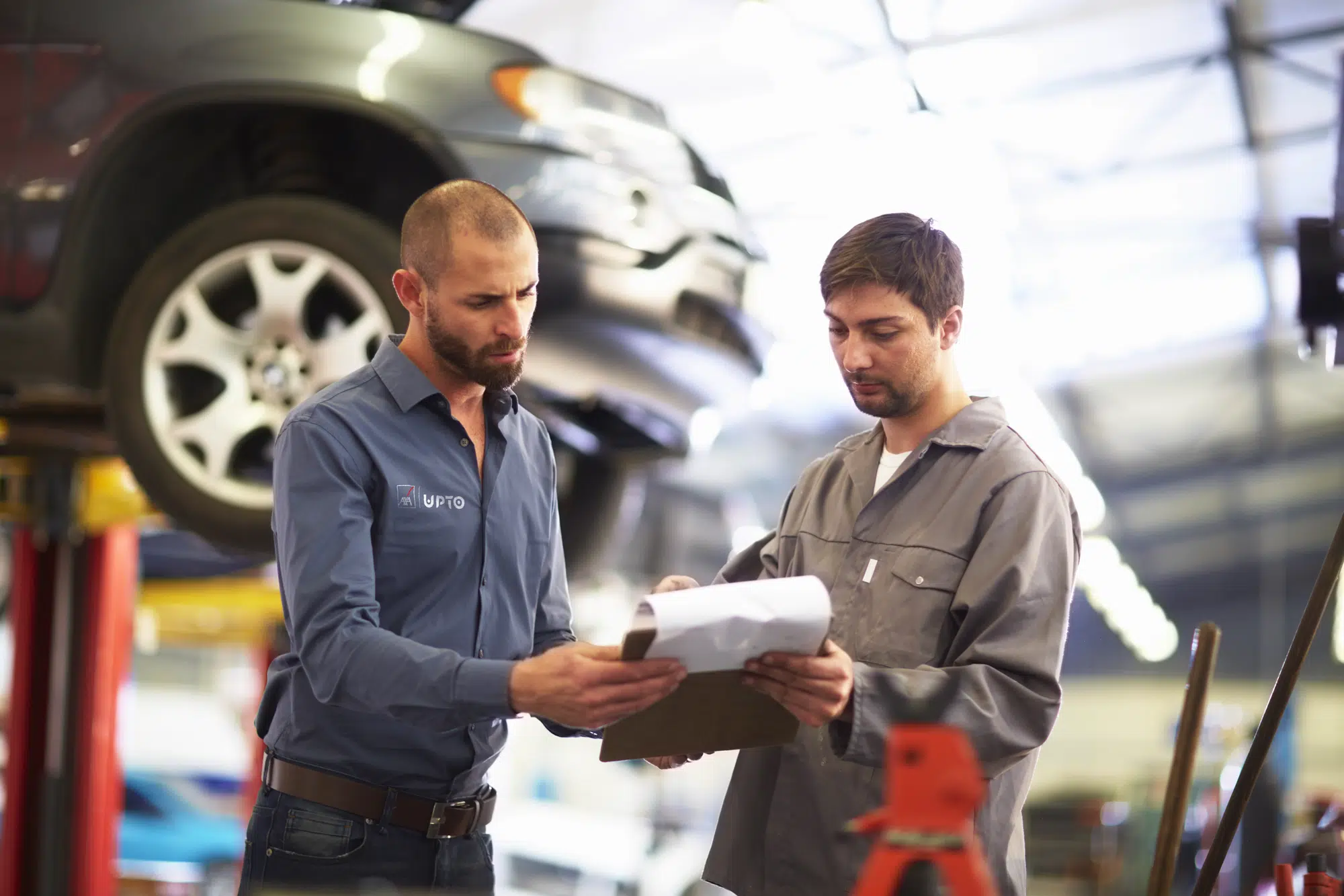 Two men in a car repair shop reviewing papers beside a lifted vehicle.