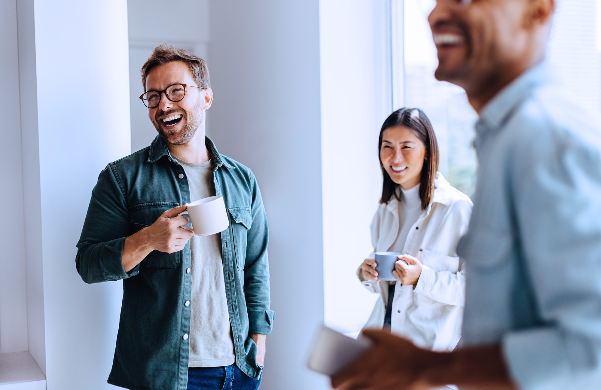 Three colleagues enjoying coffee and laughing together in a bright modern office.